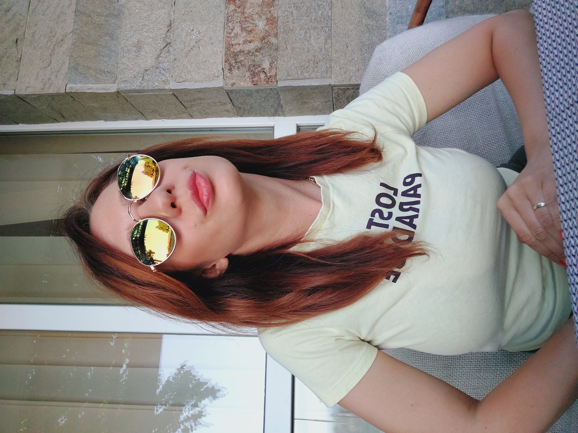 A medium shot shows a woman with reddish-brown hair and light skin wearing round, gold-rimmed sunglasses and a light yellow t-shirt with dark text that appears to read "LOST PARADISE" in reverse. She is seated and looking upwards and to her right with her lips slightly pursed. Her hands are resting on a table with a patterned surface. The background features a window with vertical blinds on the left and a stone wall on the right. The scene is likely outdoors, given the reflection of trees and a sunset in the sunglasses. The location is identified as Sozopol, Bulgaria.