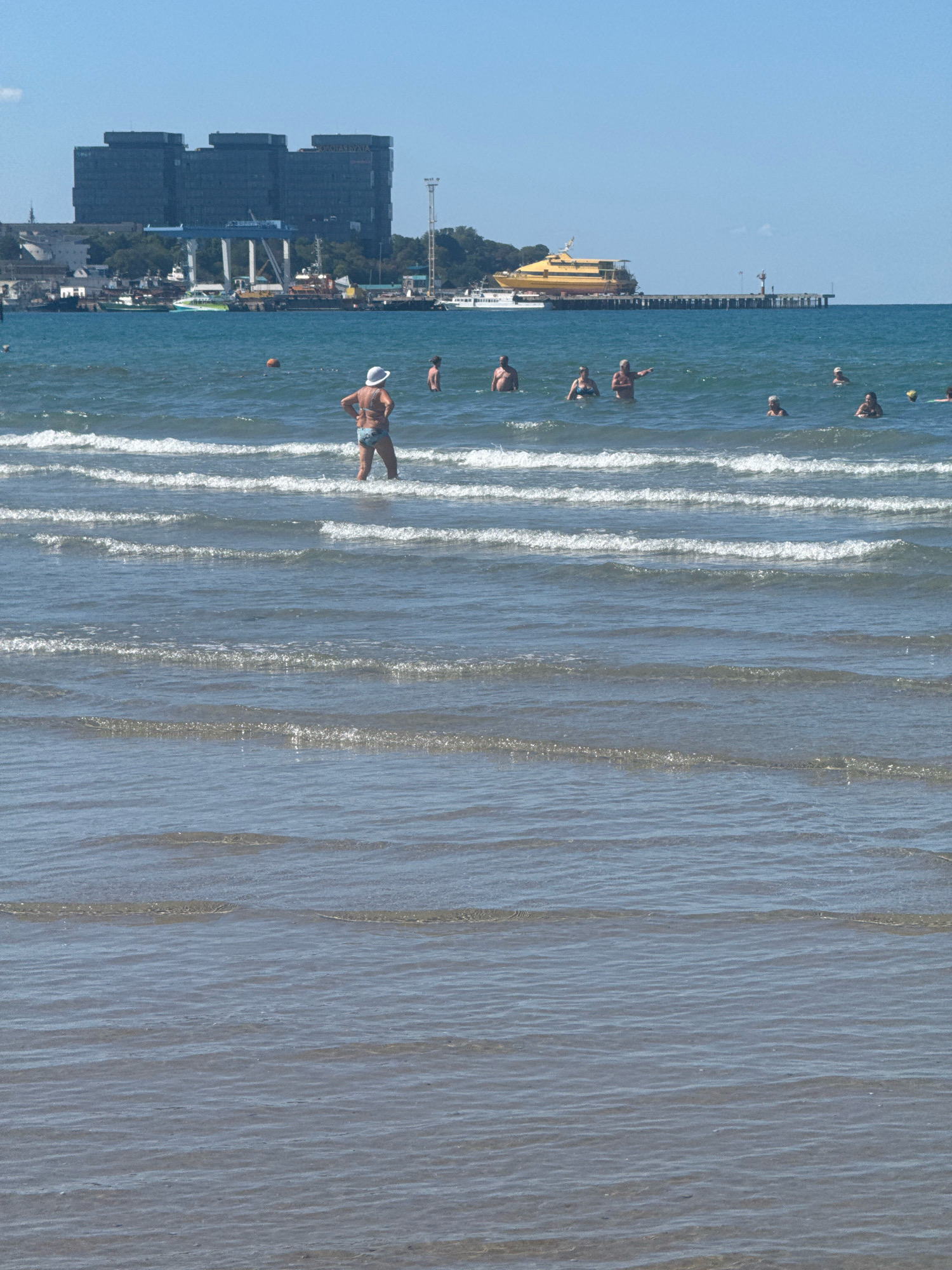 The image depicts a sunny day at a beach in Anapa, Russia. Several individuals are visible in the shallow water, some wading and others swimming. In the background, a large yellow ferry is docked at a pier, and a cluster of modern buildings dominates the skyline. The sea is calm with gentle waves rolling towards the shore. The foreground is filled with the receding waves and the sandy bottom of the sea.