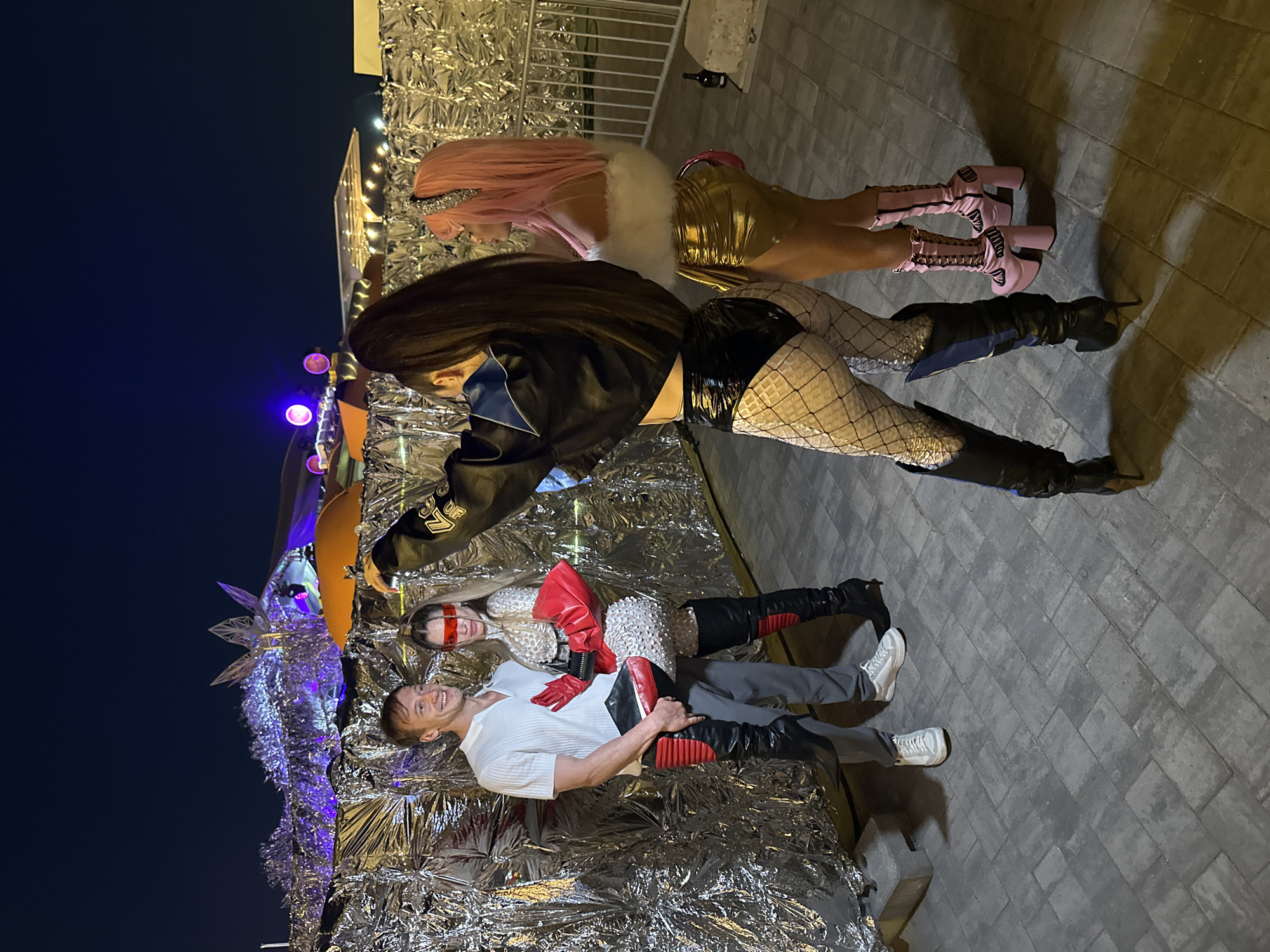 In Saint Petersburg, Russia, at night, three individuals are posing for a photograph. A man in a white polo shirt and gray pants stands to the left, with his arm around a woman dressed in a sequined outfit, red gloves, and black thigh-high boots. The woman is also wearing red blinders over her eyes. To the right of the couple, another woman with pink hair, wearing a gold mini-skirt, white fur trim top, and pink platform boots, is holding a phone and taking the picture. The background features a large, silver, metallic installation illuminated with purple lights, possibly a stage or art piece at an outdoor event. The ground is paved with grey interlocking stones, and a metal fence is visible on the right side.