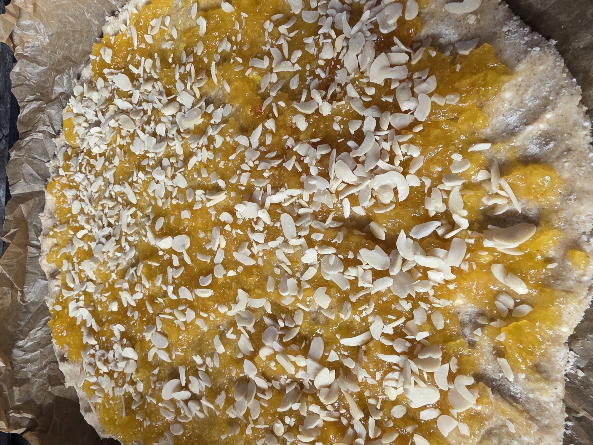 A close-up overhead shot shows a freshly baked galette with a bright orange filling, possibly apricot or peach jam, spread over a rustic, light brown crust. The crust is partially folded over the filling, creating a free-form pie shape. The galette is generously sprinkled with slivered almonds, which are scattered evenly across the top, adding texture and visual appeal. The pastry is resting on a piece of brown parchment paper, which is itself on a dark, textured surface, likely a countertop or table. The overall impression is one of a homemade, artisanal dessert, ready to be served. There is no information about the location in Saint Petersburg, Russia.