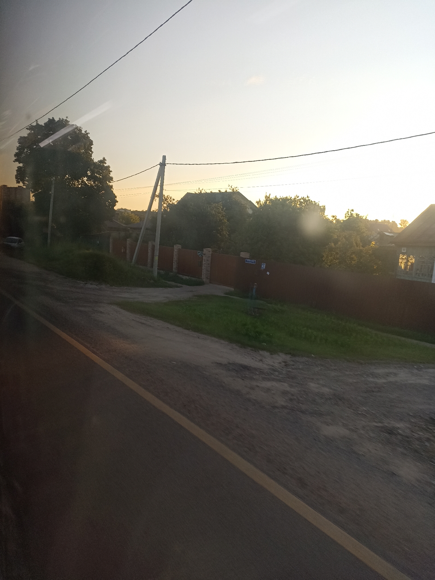 The image depicts a sun-drenched rural scene, likely taken from a moving vehicle, showing a road in the foreground with a yellow dividing line. Beyond the road, there is a grassy verge and a dark brown wooden fence. Behind the fence, trees and houses are visible. A white car is parked on the left side of the image, partially obscured by trees. Electrical poles and wires are present throughout the scene. The sky is bright, suggesting it is either sunrise or sunset. There are no people visible in the image. The image location is in Chekhov, Russia.