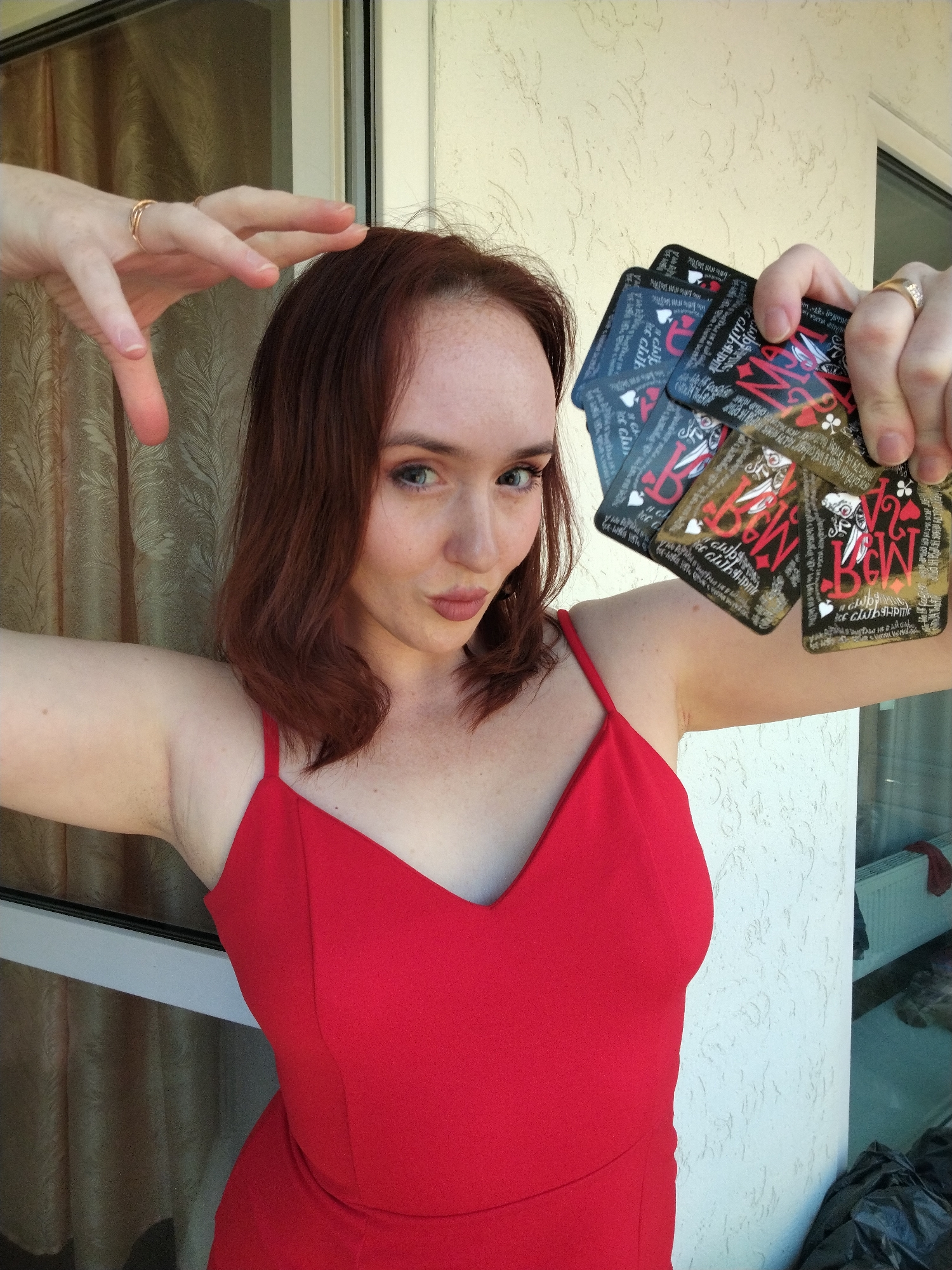 A woman with reddish-brown, wavy hair and visible freckles on her face and arms is captured in a close-up portrait, posing playfully. She wears a vibrant red, v-neck top or dress with spaghetti straps. Her blue eyes look directly at the viewer, and her lips are slightly pursed.
Her left hand is raised and bent above her head, with two simple gold rings on her ring finger. Her right hand holds a fan of several black playing cards. A gold ring with a clear stone is also visible on her right ring finger. The cards feature a black background with stylized red, white, and gold artwork, including "B&W," "club-p!nk," and various playing card suit symbols (heart, club, diamond). Intricate illustrations of what appear to be jokers or court cards are also present, along with small, stylized English text that is mostly illegible.
The background on the left consists of a textured, light brown or beige curtain. On the right, there is a textured, off-white wall, resembling stucco. A dark window or door frame runs vertically behind her, separating these background elements. In the bottom right corner, a dark object, possibly a plastic bag, and a reflection of what might be an indoor heating unit are visible through glass. The scene is brightly lit by natural daylight, suggesting an outdoor or semi-outdoor setting like a balcony or patio, located in Izhevsk, Russia.