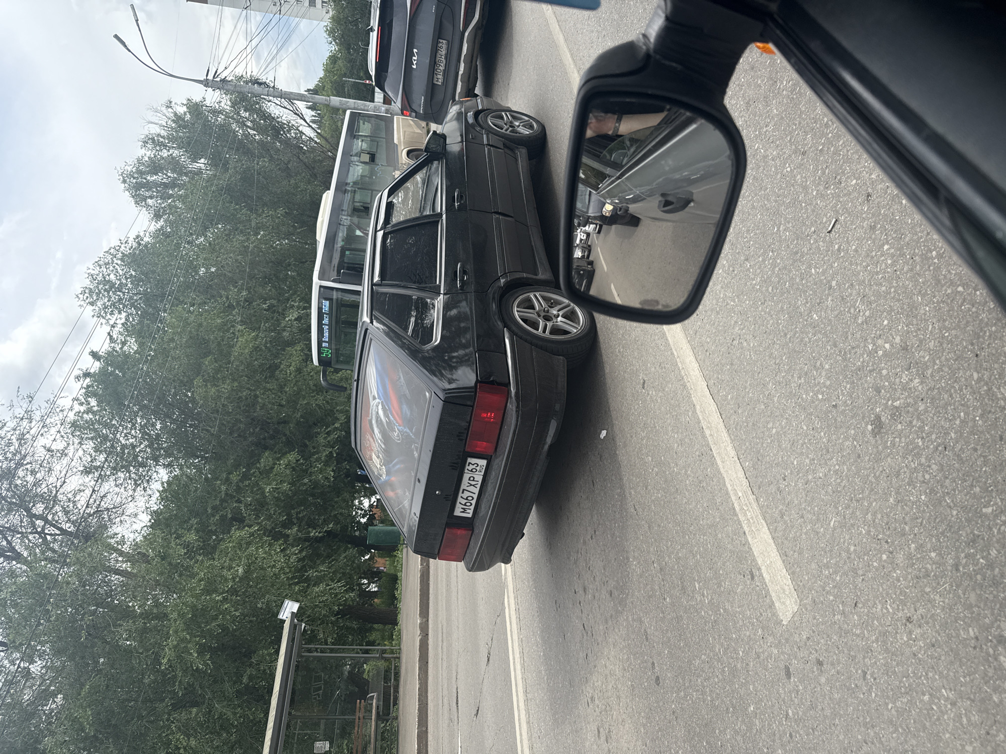 In Samara, Russia, a black, older model sedan with a distinctive decal on its rear window is seen in traffic. The license plate reads "M667XP 63 RUS". The car is positioned on a road, likely a street in the city, with trees and a bus stop visible in the background. Other vehicles, including a white bus and a dark SUV, are also present in the scene, indicating a typical urban traffic environment. The weather appears to be overcast, with a cloudy sky overhead. The image is captured from the perspective of another vehicle, suggesting the subjects are in motion.