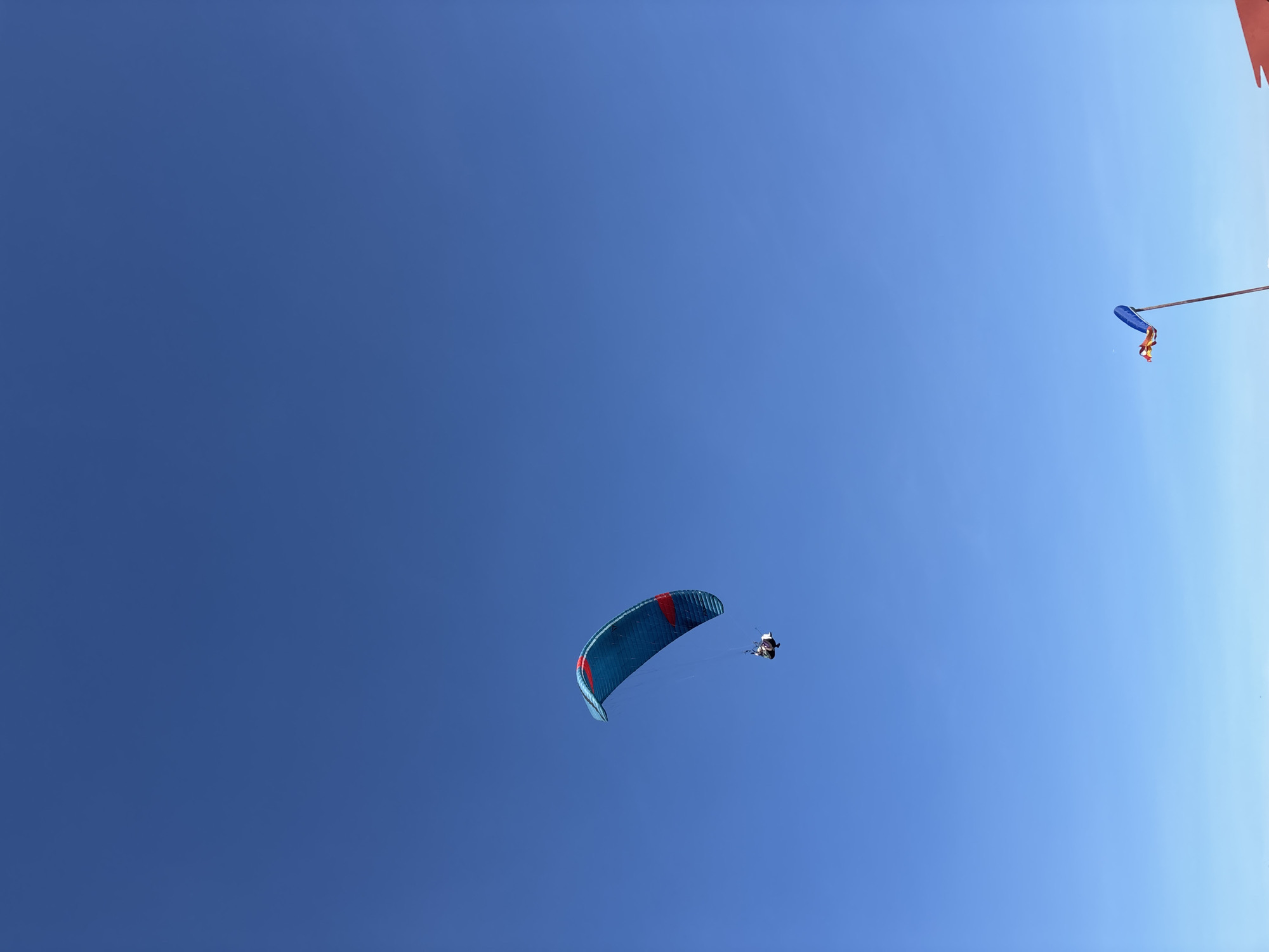 A paraglider soars in a clear blue sky, likely in Kuta Selatan, Indonesia, based on the prompt. The paraglider is blue with red accents. A person is attached to the paraglider, suspended below the canopy. In the upper right corner, there is a red and blue object hanging from a pole. The sky is clear.