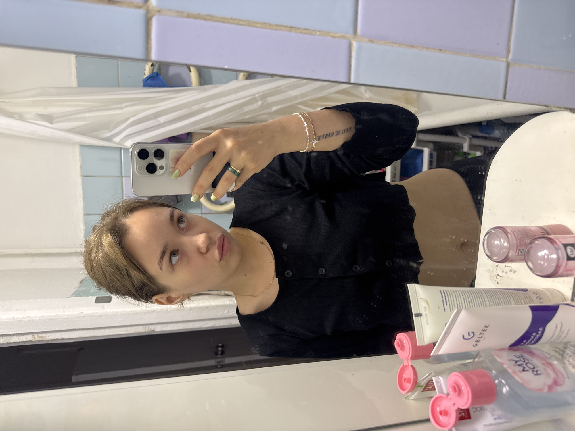 In a bathroom, a young Caucasian woman with light brown hair pulled back is taking a mirror selfie. She is wearing a black crop top and appears to be looking to her right. Her left hand holds a silver smartphone, displaying the front-facing camera. Her fingernails are painted a light green. She is also wearing a green ring on her left ring finger and a thin silver bracelet on her left wrist. A tattoo is visible on her left forearm. On the counter in front of her are several cosmetic products, including tubes of "Geltek" and "My Rose" micellar water, as well as other unidentifiable bottles and containers. The bathroom has blue and purple tiled walls, a white shower curtain, and what appears to be a white sink in the foreground. The overall environment suggests a residential bathroom.