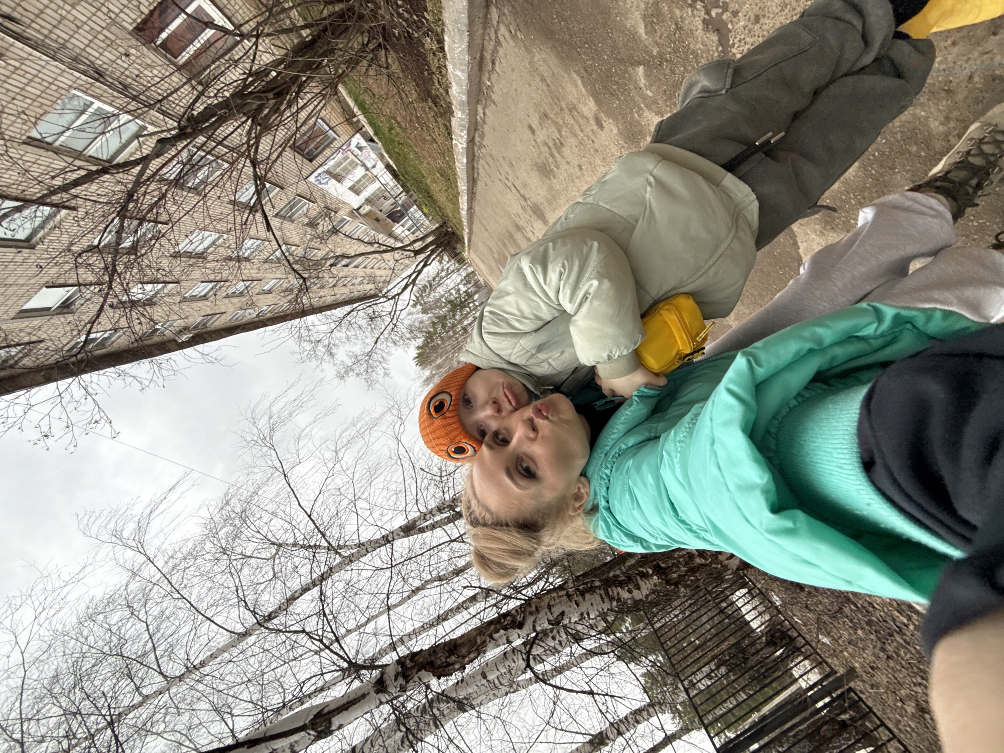 A woman and a child, both wearing winter clothing, are posing for a selfie outdoors. The woman is wearing a light green jacket, while the child has an orange hat and a light-colored jacket. The setting appears to be a park or outdoor area, with bare trees visible in the background. A multi-story brick building is also visible. Based on the metadata, the location is Chaikovsky, Russia.