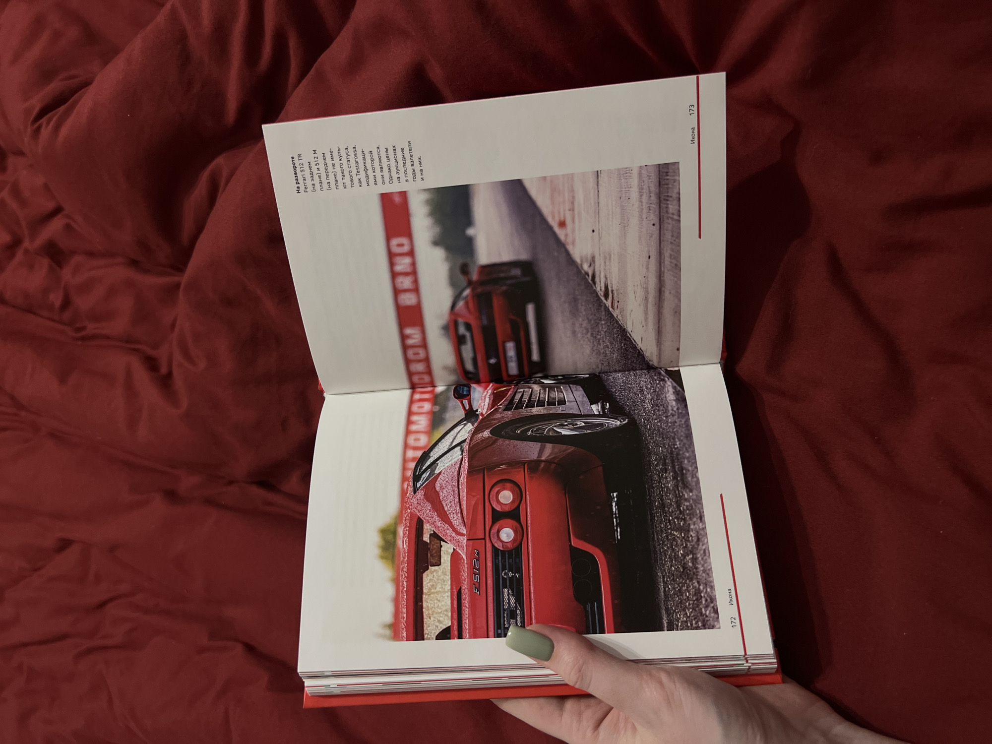 The image shows an open book resting on a red textured surface, likely a bed or blanket. The left page features a close-up rear view of a red Ferrari Testarossa on a wet road. The right page displays text and a photograph of two red Ferraris on a race track with a banner reading "AUTOMOTODROM BRNO". The text on the right page appears to be in Russian, discussing Ferrari models. A person's hand with light green painted fingernails is visible on the left side of the book, partially obscuring the left page. The pages are numbered 172 and 173 at the bottom. There is no indication of the location being in Дачное, Russia.