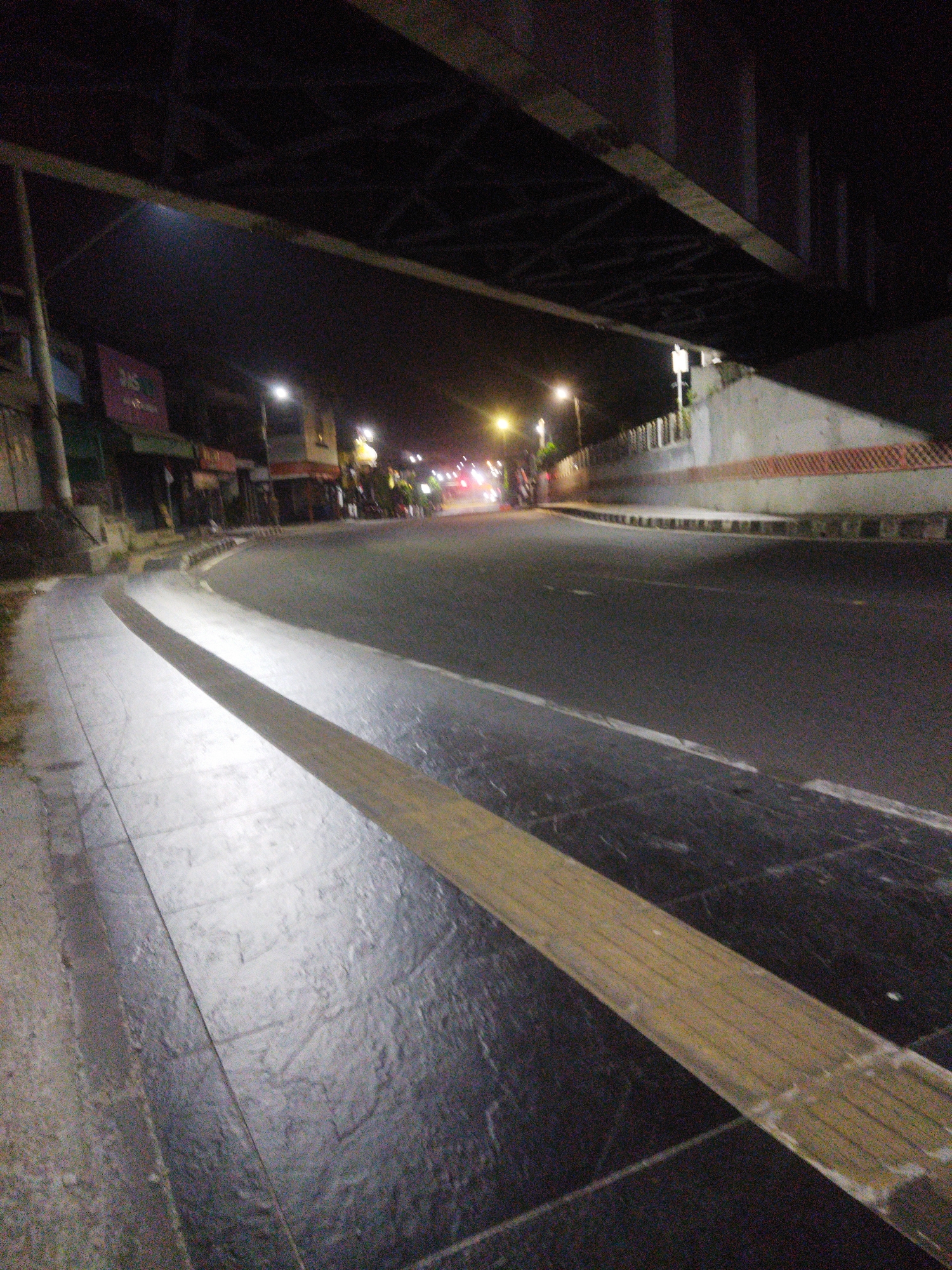 The image depicts a nighttime view of a city street in Wonogiri, Indonesia, taken from the perspective of a sidewalk. An overhead structure, possibly a bridge or overpass, dominates the upper portion of the frame. To the left, shops are visible with illuminated signs. The street itself is paved and curves to the right, with a distinct yellow tactile paving strip running along the edge of the sidewalk. Streetlights illuminate the scene, casting reflections on the wet pavement. The overall atmosphere is dark and urban, typical of a city at night. No individuals are clearly discernible in the image.