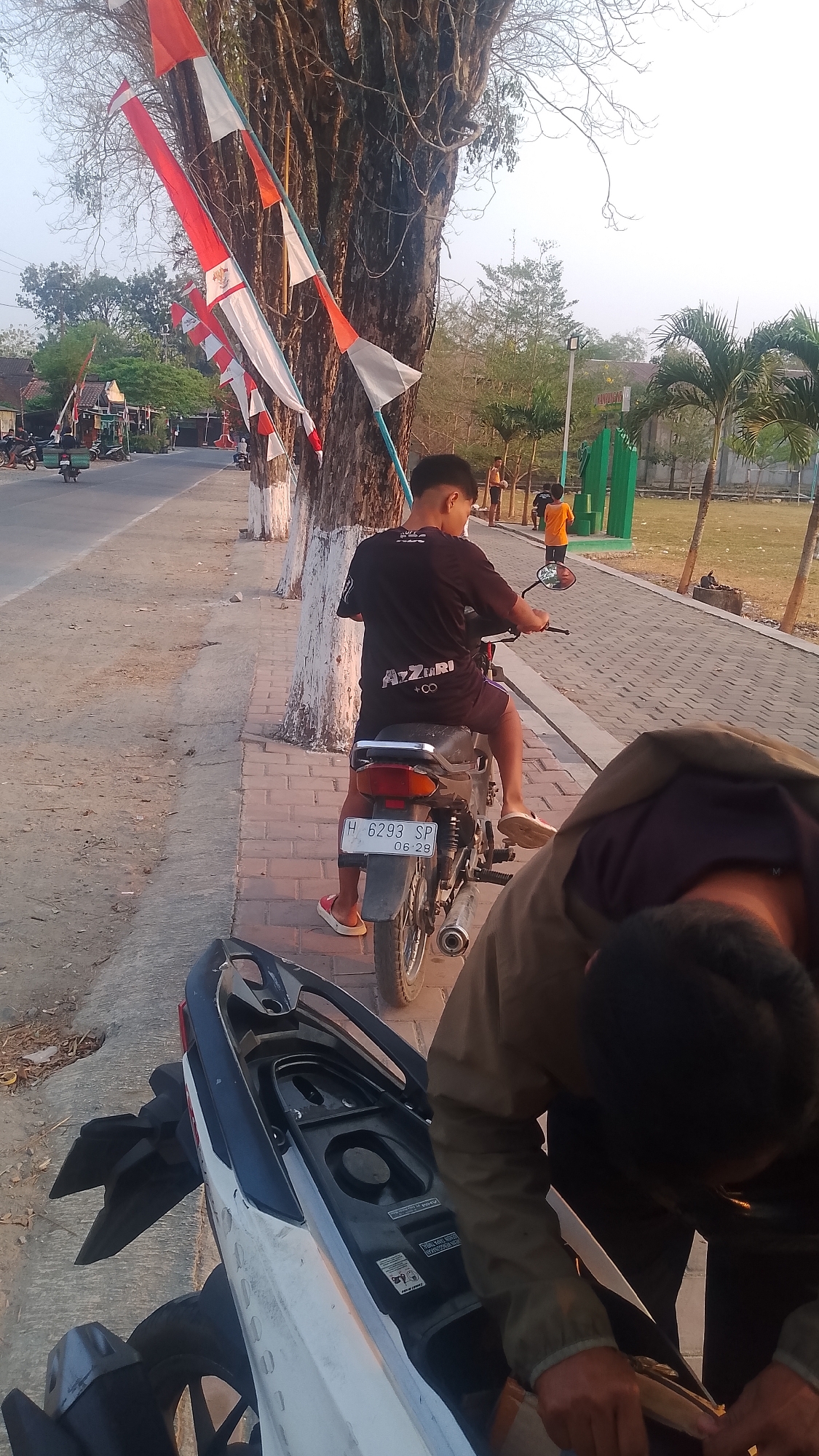 In Karanglangu, Indonesia, a young male sits astride a black motorcycle parked on a brick walkway. He wears a black t-shirt with white lettering and dark shorts, his feet clad in red sandals. The motorcycle's license plate reads "H 6293 SP 06-29." To the right, another individual, seen from behind, is bent over, appearing to adjust something on a white scooter. A large tree with white-painted sections stands to the left, adorned with red and white Indonesian flags. In the background, a paved road is visible with scattered parked motorcycles and a few people walking. Palm trees and buildings can be seen further in the distance under a hazy sky.