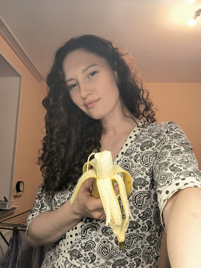 A mid-shot selfie captures a woman indoors, within what is identified as a location in Saint Petersburg, Russia. The subject is a female with long, dark, curly hair, looking directly at the camera with a slight smile. She wears a light-colored garment patterned with black paisley and polka dots. In her left hand, she holds a yellow banana, partially peeled with approximately two-thirds of the peel drawn down, revealing the fruit. The tip of the banana appears bitten or broken off. The background consists of light peach-colored walls and a white ceiling. A ceiling light fixture with two exposed bulbs is visible in the upper right, emitting warm illumination. To the left, a black circular object with a white face, possibly a clock or thermostat, is mounted on the wall, with a white plastic basket on a shelf below it. A metallic clothes drying rack, holding dark fabric, is partially visible in the lower-left corner of the frame.