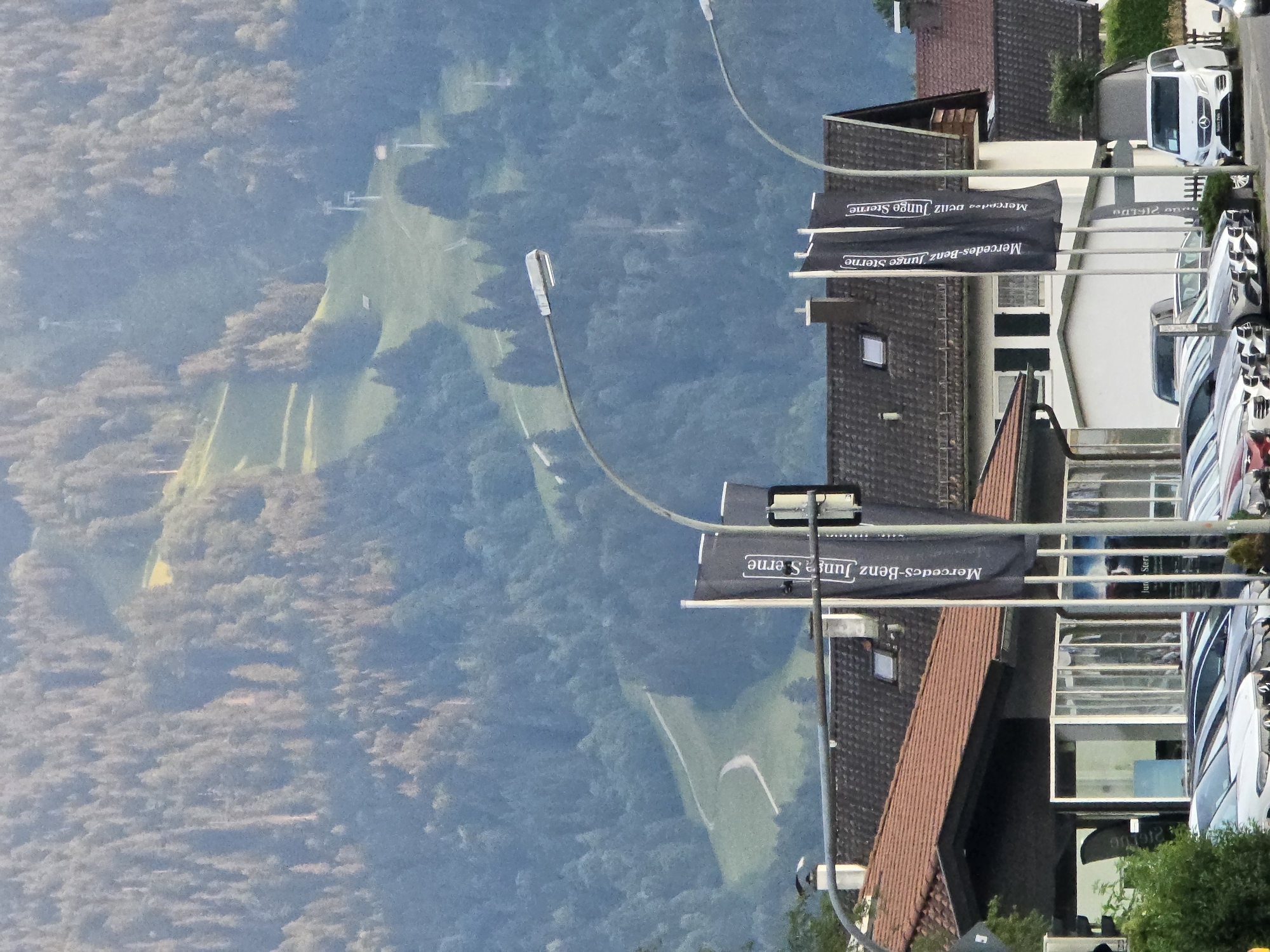 The image depicts a daytime scene in Garmisch-Partenkirchen, Germany. In the foreground, a Mercedes-Benz dealership is visible, with several cars parked outside and two black flags with white lettering that reads "Mercedes-Benz Junge Sterne" (Mercedes-Benz Young Stars) displayed prominently. The dealership has large glass windows. In the middle ground, a residential or commercial building with a dark tiled roof is situated behind the dealership. Further in the background, a tree-covered mountain slope dominates the view, with a ski slope clearly visible on its upper half. Several ski lift poles and cables are scattered across the mountainside, indicating it's a ski resort area. The overall atmosphere suggests a clear day with soft lighting, likely during the late afternoon or early morning. There are no people clearly visible in the image.