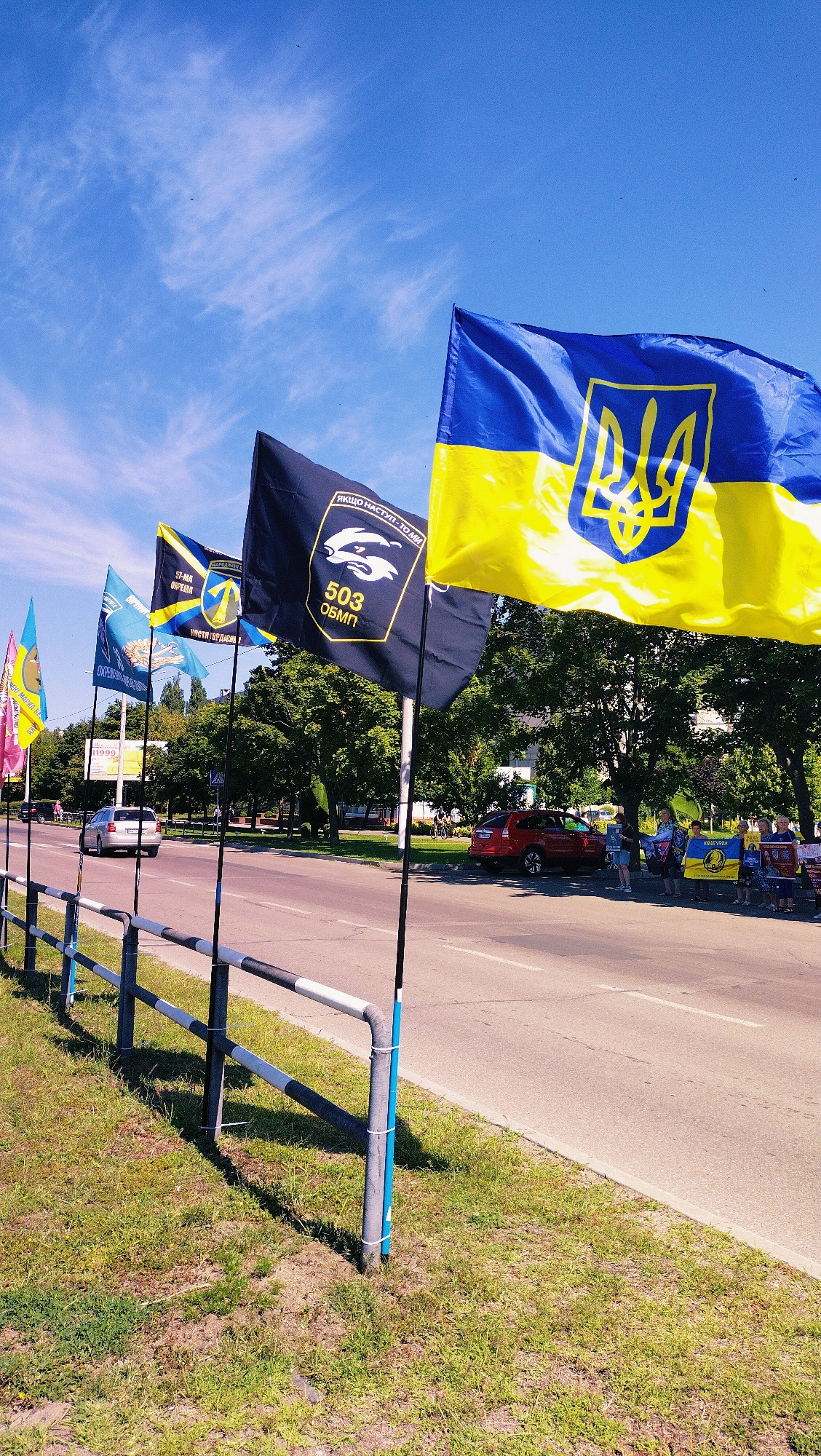 In Horishni Plavni, Ukraine, on a sunny day, several flags are displayed along a roadside grassy area. The prominent flag in the foreground is the national flag of Ukraine, a horizontal bicolor of blue on top and yellow on the bottom, featuring the Ukrainian trident in the center. To the left of the Ukrainian flag, a dark flag displays a white emblem and text that appears to read "503 ОБМП". Further to the left, other flags are visible, including one with a yellow trident on a blue background and a flag with vertical stripes of yellow, blue, and white. A metal railing with black and white stripes separates the grassy area from the road. Cars are present on the asphalt road, including a silver sedan and a red SUV. In the background, trees line the street, and several individuals are gathered, some holding flags. The sky is clear blue with scattered white clouds.