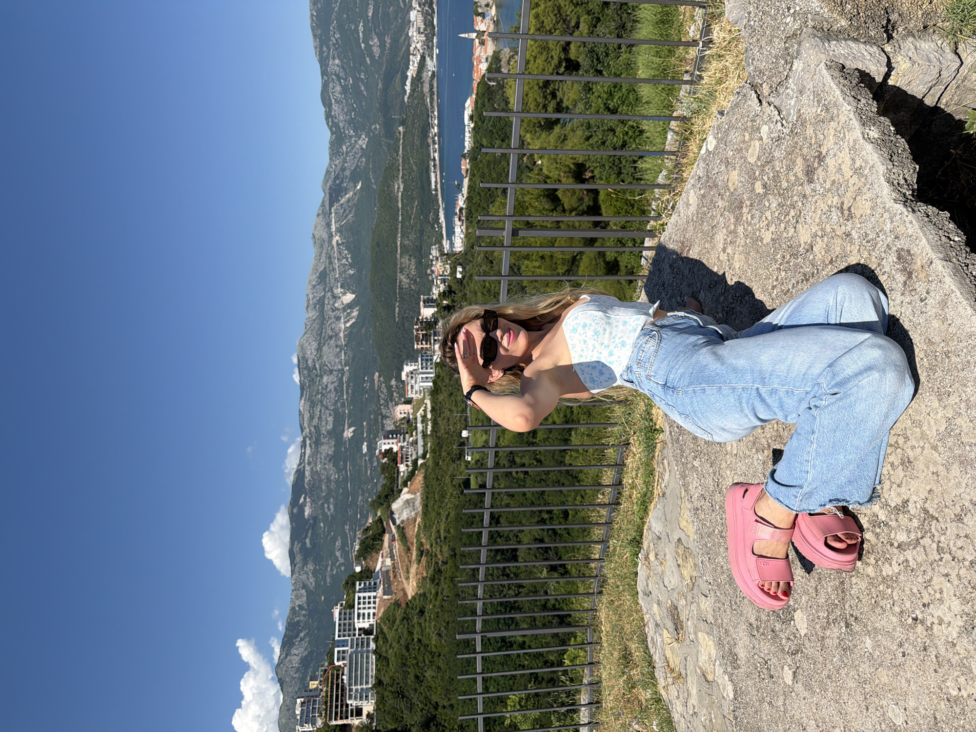 A young woman with blonde hair and sunglasses is depicted kneeling on a light grey stone surface under a clear blue sky. She wears a white and light blue floral corset top, light blue wide-leg jeans, and pink platform sandals, revealing red nail polish on her toes. Her right hand shields her eyes from the bright sunlight as she looks towards the camera with a slight smile.
Behind her, a dark grey metal fence spans horizontally. Beyond the fence, a sloping terrain is covered with dense green trees. In the mid-distance to the right, the coastal town of Budva, Montenegro, is visible, featuring numerous white and light-colored buildings with red roofs clustered along the coastline. A prominent church tower, characteristic of Budva's Old Town, rises above the buildings near the water's edge. A calm, deep blue bay with several boats extends into the background. On the left side, modern buildings and construction sites are situated on a green hillside. A range of large, rugged mountains, predominantly green with some exposed rock, forms the distant horizon. The scene is brightly illuminated by direct sunlight, casting distinct shadows.