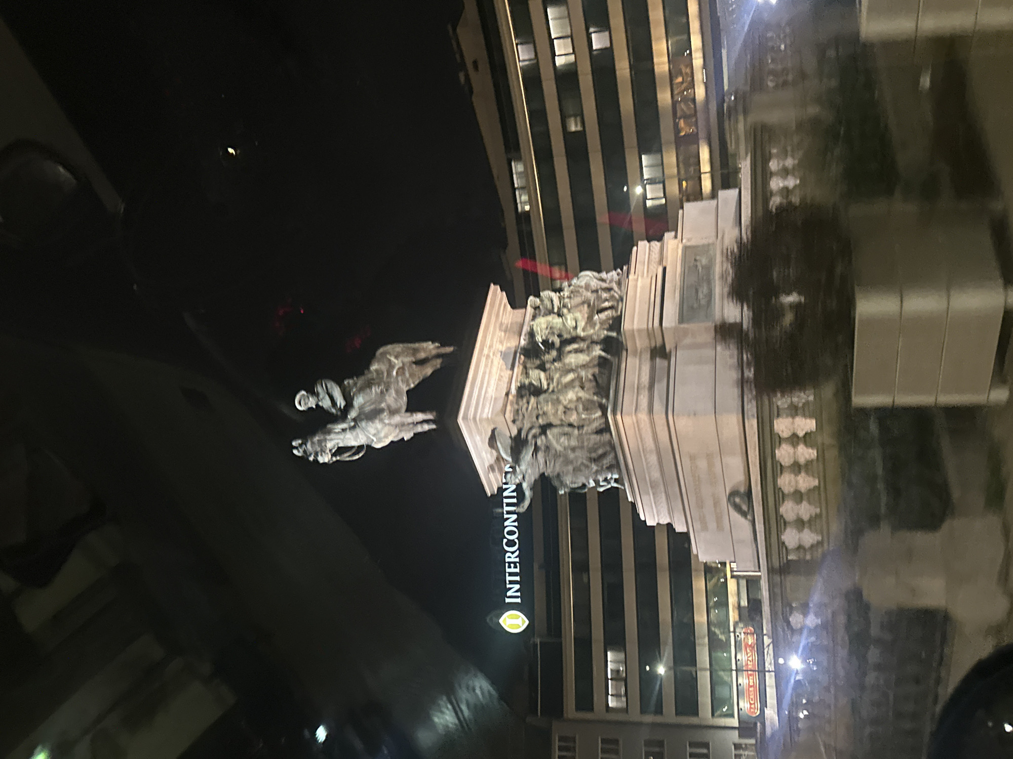 The image displays the Monument to Tsar Liberator, an equestrian statue located at pl. Narodno Sabranie in Sofia, Bulgaria, captured at night. The bronze statue of a man on horseback is prominently positioned atop a large, multi-tiered light-colored granite pedestal adorned with numerous bronze reliefs depicting figures. In the background, the InterContinental Sofia hotel is identifiable by its brightly illuminated "INTERCONTINENTAL" sign and logo on a multi-story building featuring multiple illuminated windows. Another large building with visible illuminated windows stands to the monument's right. The immediate foreground includes a low balustrade-style wall and a rectangular planter box, partially obscured by reflections on a reflective surface. No individuals are visible.