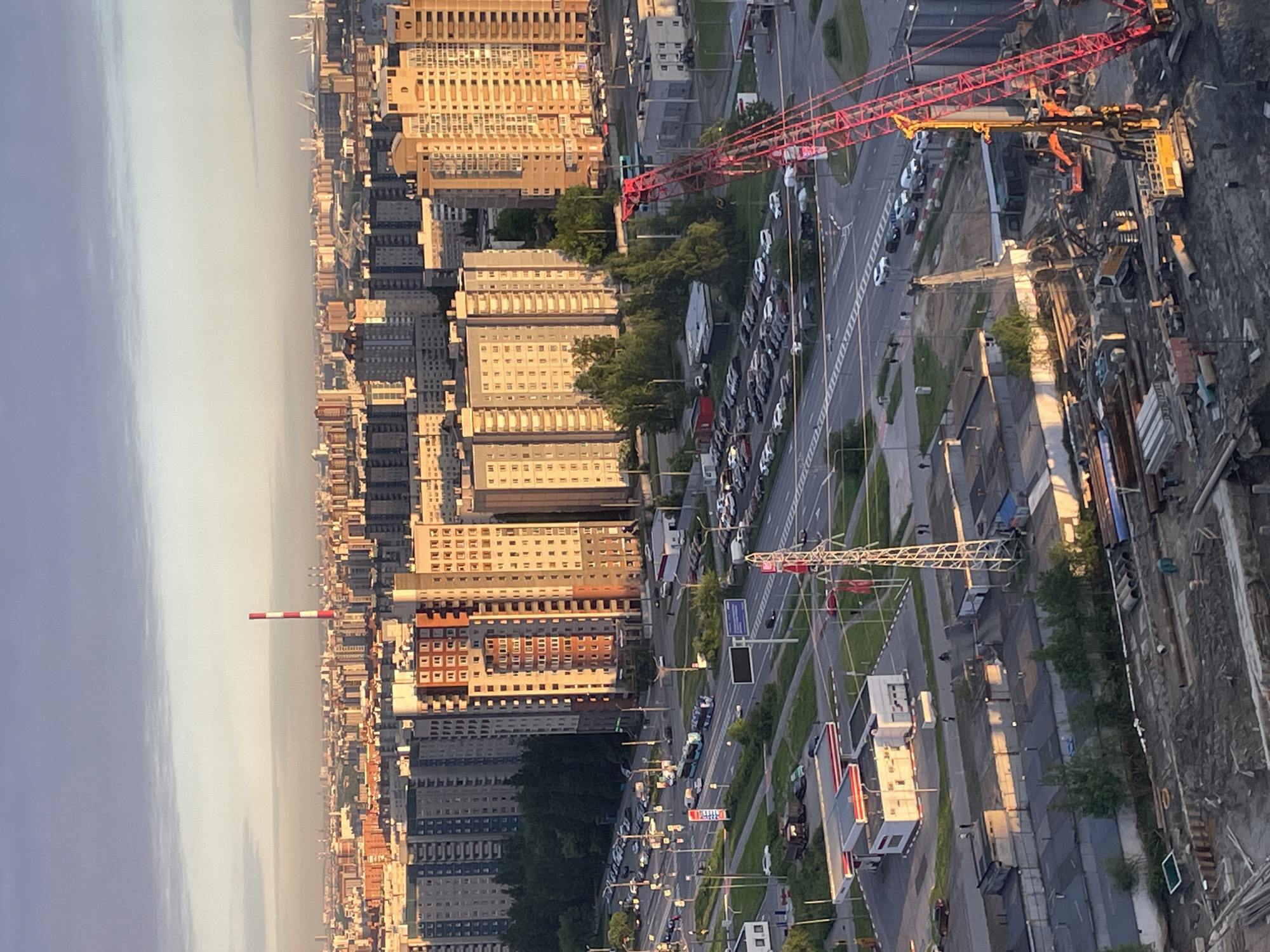 This high-angle, panoramic photograph captures an expansive urban landscape, likely in Saint Petersburg, Russia, during the late afternoon or early evening. The scene is illuminated by warm, golden sunlight, casting long shadows across the city and highlighting the facades of numerous buildings.
The cityscape is dominated by multi-story residential or commercial buildings, many displaying beige, grey, and some reddish-brown exteriors typical of post-war or Soviet-era architecture. These structures stretch far into the hazy distance, creating a dense urban fabric. A wide, multi-lane road traverses the scene, with vehicles parked along its shoulders and moderate traffic visible. Streetlights, power lines, and road markings line the thoroughfare.
Prominently featured in the right foreground and mid-ground is a large red and white tower crane, signifying active construction. Adjacent to the road, a construction site is visible, filled with excavated earth, various pieces of heavy machinery, and partially built elements. Further along the road, another, smaller white construction crane can be seen. Pockets of green trees and bushes are interspersed throughout the urban environment, primarily along the road and between buildings.
In the far distance on the left side of the frame, a distinctive red and white striped industrial chimney or stack stands tall against the horizon. The sky above transitions from a lighter, somewhat hazy tone near the horizon to a deeper, clearer blue-grey overhead. Although several signs are visible on buildings and along the road, any text on them is unreadable due to the distance and image resolution. The overall impression is of a vast, developing city during a tranquil hour of the day.