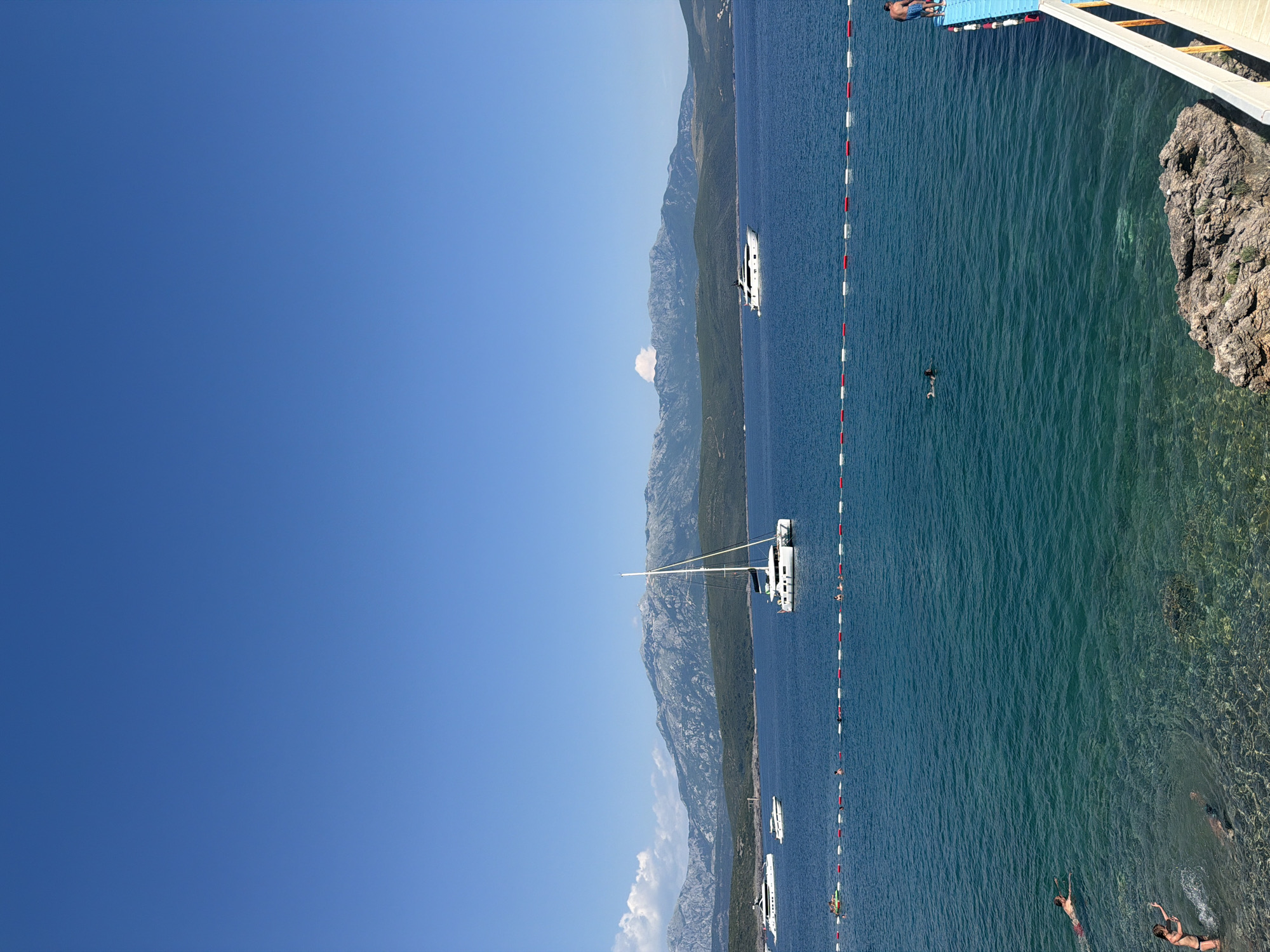 The image depicts a scenic coastal view on a clear, sunny day, likely in or near Radovići, Montenegro. The turquoise waters of the sea dominate the foreground, with several people swimming and enjoying the water. The seabed is visible in some areas due to the clear water. A roped-off swimming area, marked by buoys with red and white stripes, separates the shore from deeper water.
Further out in the bay, various boats are anchored. Prominently featured is a large white catamaran with a tall mast, and several other yachts and smaller vessels are scattered across the water. In the background, a range of mountains, some with rocky outcrops and covered in green vegetation, forms a dramatic backdrop against the bright blue sky.
On the right side of the image, a blue pontoon dock is visible, with a person standing on it. The overall scene suggests a popular tourist or recreational area.