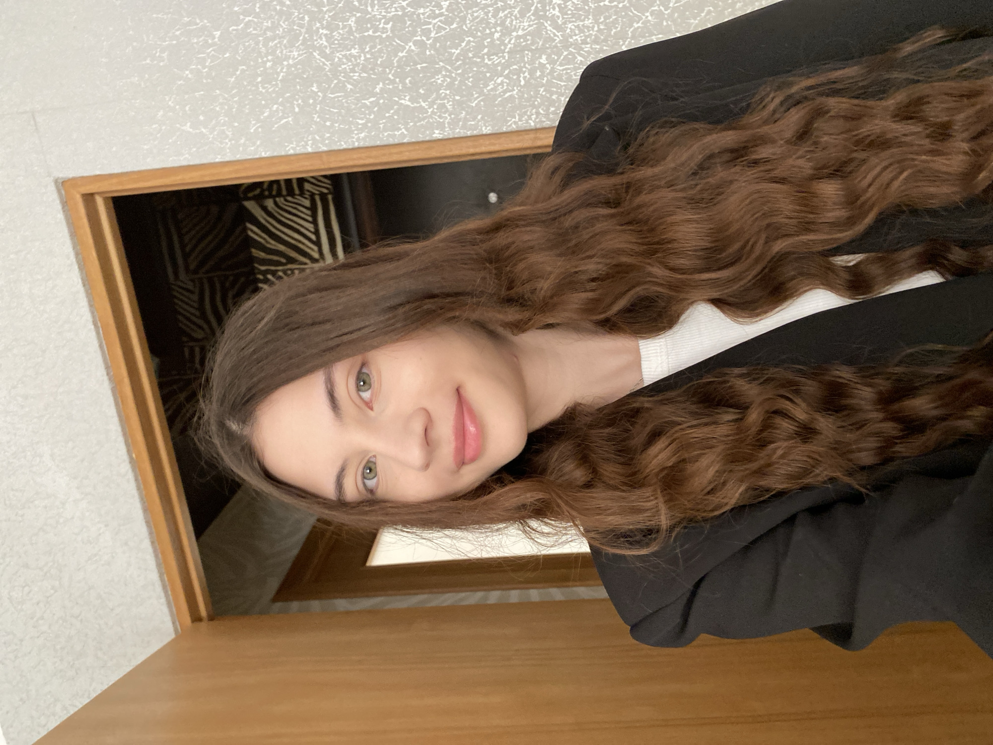 A young woman with long, wavy brown hair is visible from the shoulders up. She is wearing a white shirt under a black blazer. Her face is angled towards the viewer, and she is smiling slightly. The background features a partially visible door frame and a wall with a textured pattern. Inside the door frame, there is a glimpse of a patterned wall covering. The location cannot be determined as Votkinsk, Russia, since there is no information visible in the image to indicate the specific location.