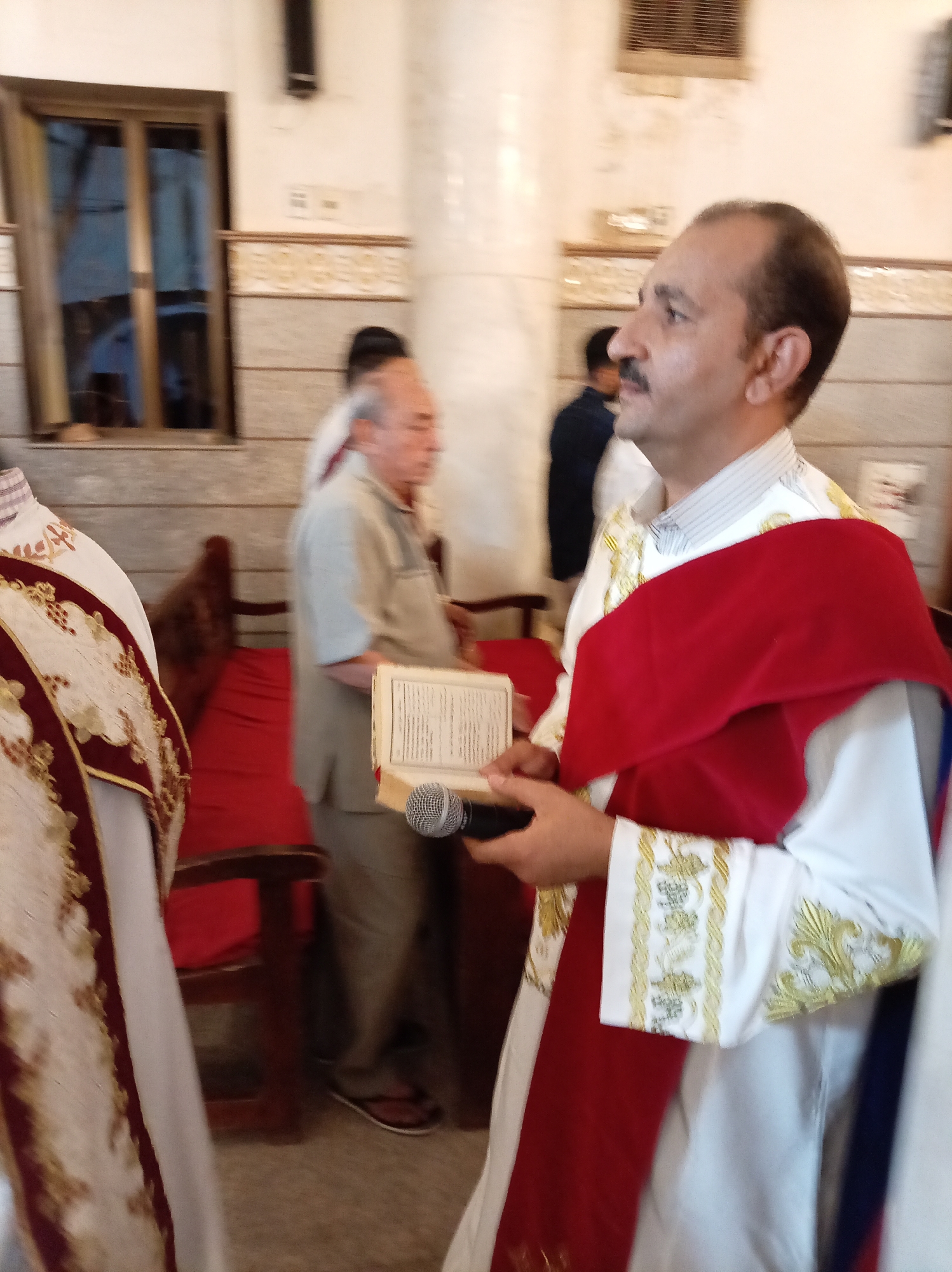 A medium shot reveals a male in a white robe with gold detailing, draped with a red cloth over his shoulders, holding a book and a microphone. He is standing in what appears to be a church or religious setting. Behind him, a man in a tan shirt and pants stands near a red upholstered bench. A window is visible in the background. The location appears to be the city of Unknown, Unknown.