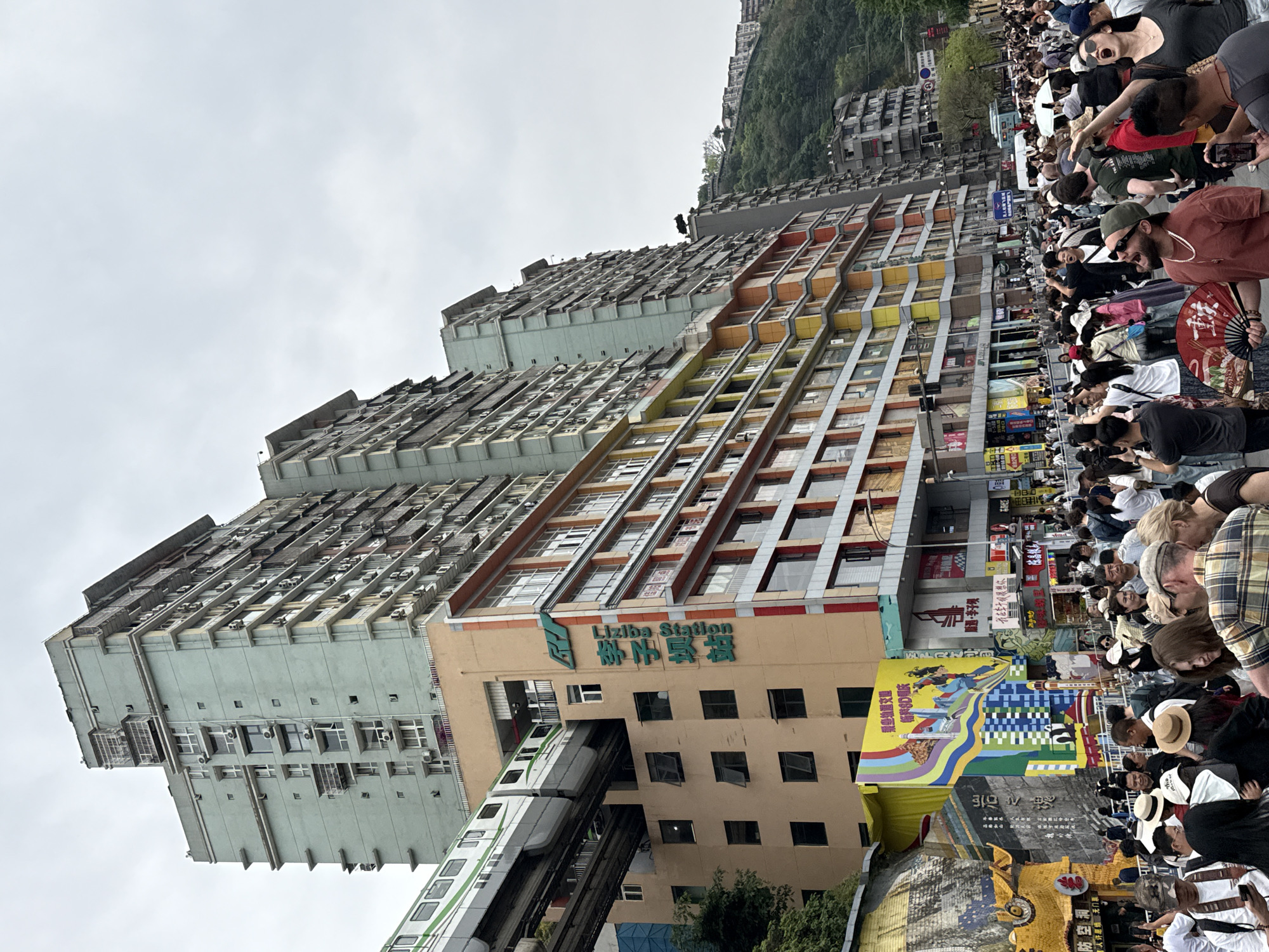 Here is a description of the image.
The image depicts a bustling street scene in Yuzhong District, China, specifically at the Liziba Station. In the foreground, a large crowd of people is visible, many of whom are taking photos with their phones or cameras. The focus of the crowd's attention is likely the Liziba Station, a building with a modern train passing through its structure. The building is painted in a combination of beige and gray. Colorful banners and advertisements adorn the lower levels of the building. In the background, high-rise apartment buildings stretch into the sky, creating a dense urban landscape. The sky is overcast, providing a neutral backdrop to the scene. The image captures the unique architectural feature of a train passing through a building, a landmark that attracts many visitors.