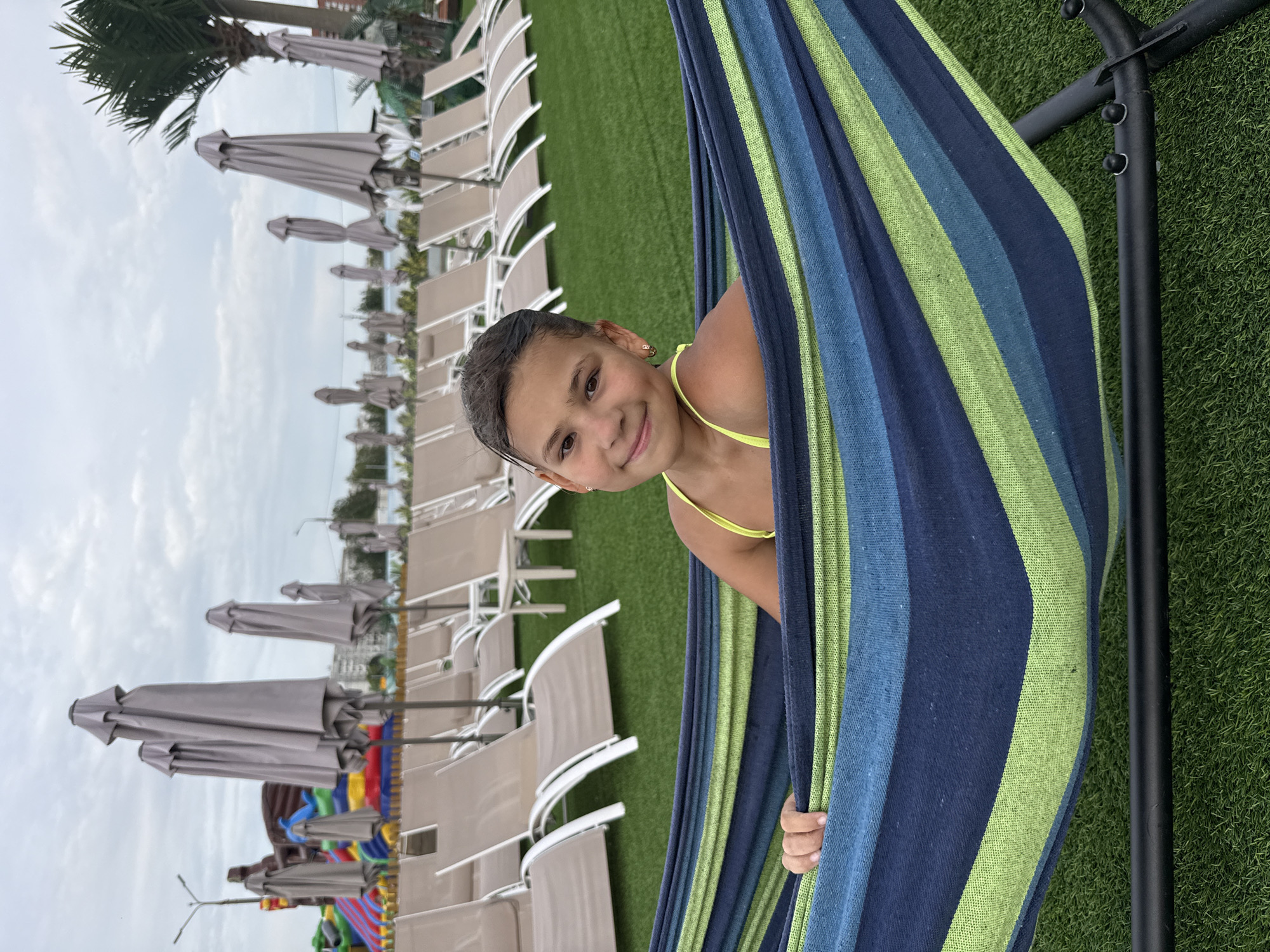 A young girl with dark hair and a bright yellow swimsuit peeks out from a striped hammock. The hammock, made of blue and green fabric, is supported by a black metal frame. She is smiling at the camera, her expression conveying happiness and relaxation. The background shows a neatly manicured green lawn, and beyond that, rows of closed, light-colored beach umbrellas and lounge chairs. The sky appears overcast, suggesting a cloudy day. The overall scene depicts a leisure setting, possibly a resort or a park, with a focus on a child enjoying a relaxing moment outdoors. The location is identified as Rusanovo, Russia.