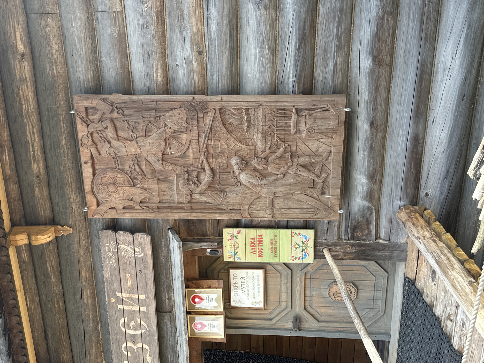 The image shows the exterior of a wooden log building, likely a museum or shop, in Nevskii Parkleskhoz, Russia. A large, intricately carved wooden panel depicting a blacksmithing scene hangs on the wall. In the panel, a blacksmith is shown hammering metal, with a young apprentice assisting. There are also signs on the building. One sign, in Cyrillic script, reads "ОТКРЫТО МУЗЕЙ" which translates to "Museum OPEN." Another sign below it states "УСАДЬБА "БОГОСЛОВКА"" meaning "Estate 'Bogoslovka'." There is also a sign with a phone number. To the right of the entrance, a vertical sign with painted birds and floral designs reads "ЛАВКА КОСТИНА" which translates to "Kostina's Shop." Below that, it says "СУВЕНИРЫ РУЧНОЙ РАБОТЫ" meaning "Handmade Souvenirs." A wooden ramp leads up to the entrance, which has a traditional wooden door. The overall impression is one of traditional Russian architecture and craftsmanship.