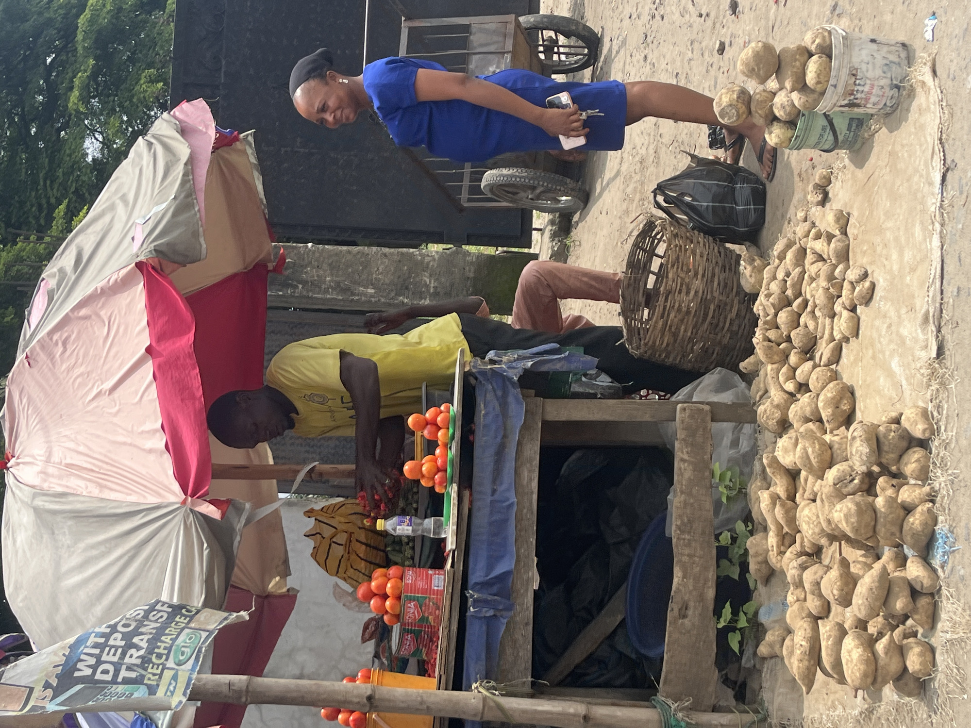 An outdoor market stall in Ado, Nigeria, displays a variety of produce. A man in a yellow shirt stands behind a wooden table, arranging tomatoes. Piles of tomatoes are visible on the table and in a yellow bucket on the left. To the right, a woman in a blue dress stands, holding a phone and keys. In the foreground, a large mound of yams is piled on the ground, with two buckets also filled with yams to the right. A woven basket and a black bag are placed near the woman. Behind the stall, a metal cart with wheels is visible. The ground is a mixture of dirt and sand. The scene is shaded by a large umbrella with pink, grey, and red panels. A sign on a bamboo pole on the left advertises "WITH DEPOSIT TRANSFER RECHARGE CARD."