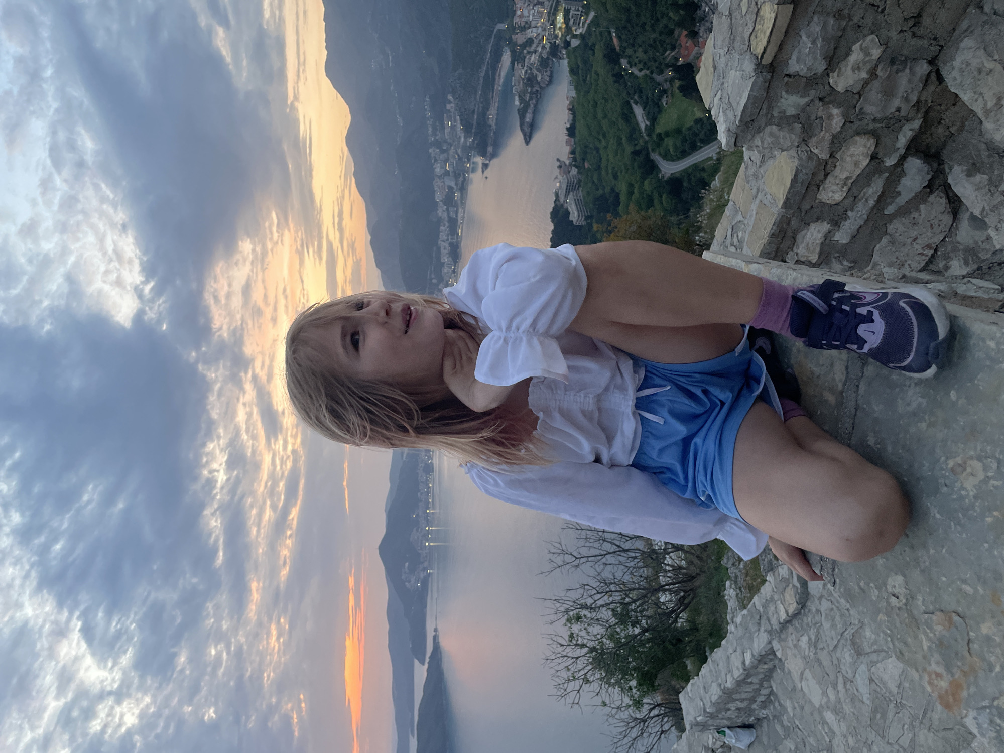 A young child, likely a girl with light-colored hair, is captured in a scenic image, sitting on a stone ledge or wall. She is wearing a white long-sleeved top with ruffled cuffs, blue shorts, and dark athletic shoes with pink socks. Her knees are bent, and she leans on one arm, looking upwards and slightly to her left with a serene or contemplative expression.
The setting is a picturesque coastal landscape, identifiable as Sveti Stefan, Montenegro. Behind the child, a sprawling bay or sea extends into the distance, reflecting the ambient light. Along the irregular coastline, a town or city is visible, with buildings dotting the shore and lights beginning to twinkle, indicating either dusk or dawn. Towering mountains frame the bay in the background. The sky above is adorned with scattered clouds, displaying a gradient of colors from warm golden and orange hues near the horizon to cooler blues and grays higher up, typical of a twilight hour. Sparse vegetation, including trees, can be seen on the slopes leading down to the water and near the stone structure where the child sits. The image captures a moment of quiet appreciation for the dramatic view from a high vantage point. There is no visible text.