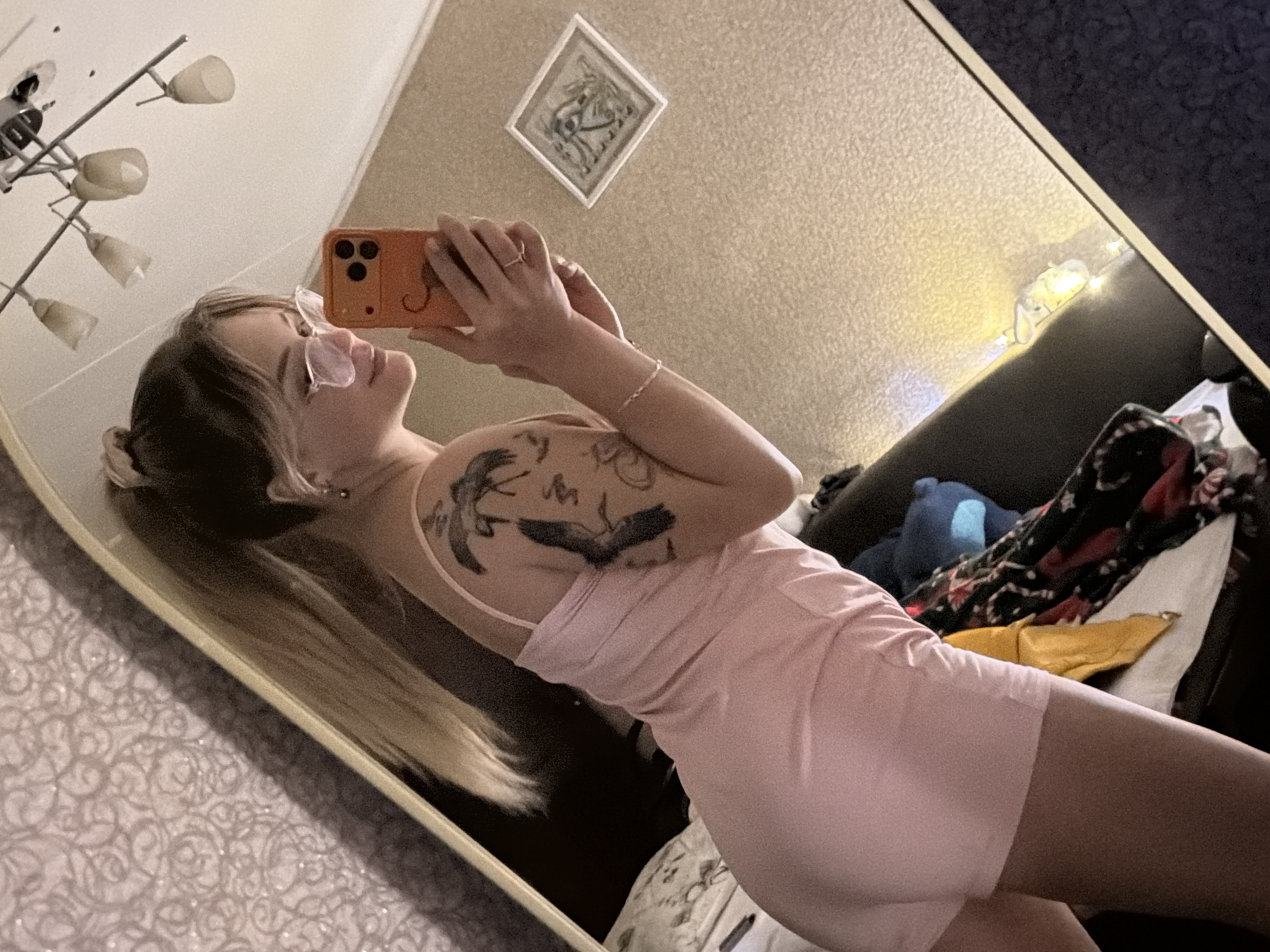 A young woman is taking a selfie in a mirror. She is wearing a pink dress and glasses. The location is likely an apartment in Naberezhnye Chelny, Russia. Evidence for this includes the typical apartment decor and layout, and the style of light fixtures and wall hangings common in Russian homes. The woman is holding an orange-colored smartphone. She is partially visible in the mirror. The mirror also reflects part of a room, with a sofa, a pillow and blankets. The lighting suggests it is evening or a poorly lit room.