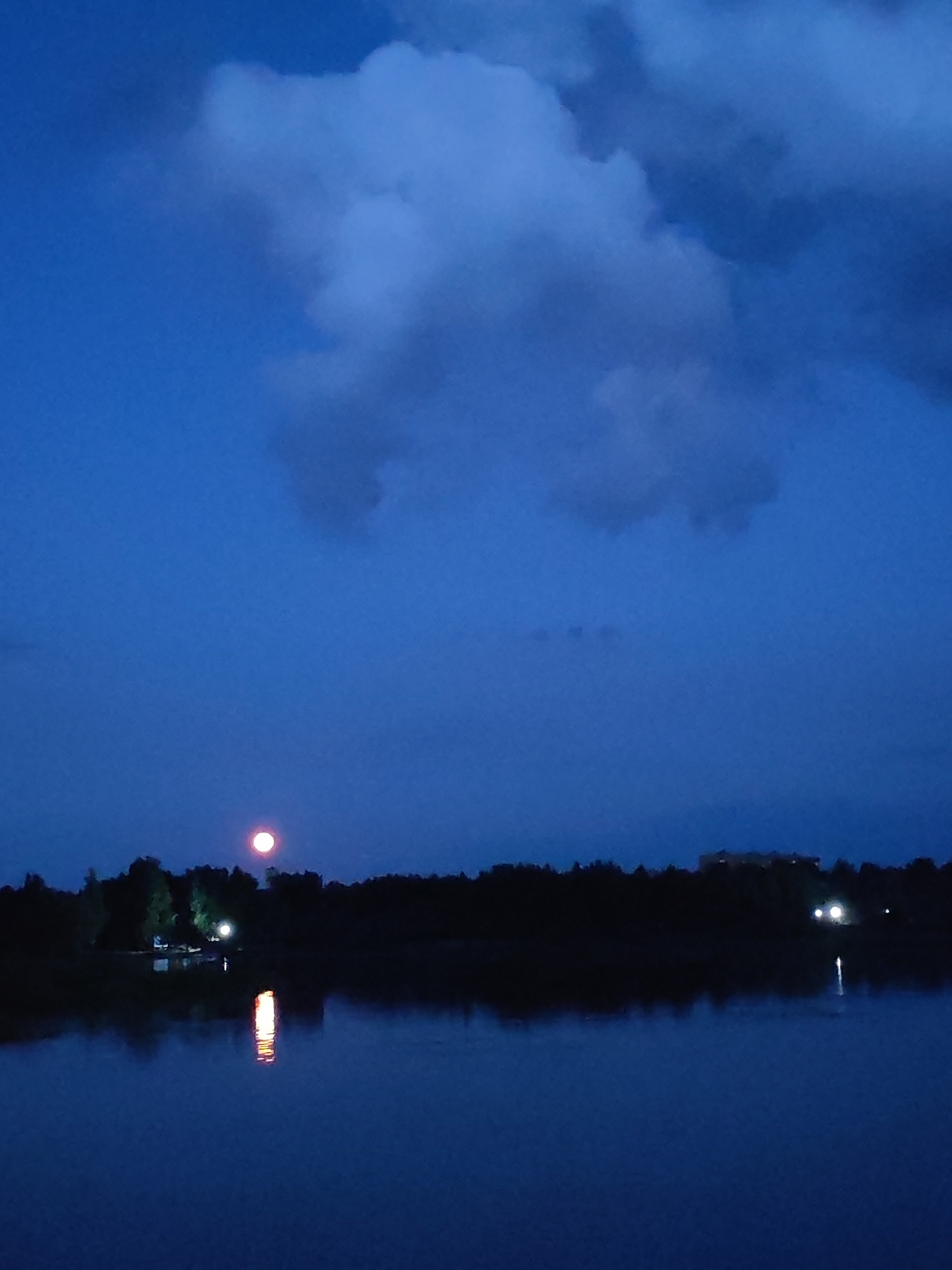 The image depicts a twilight scene over a body of water, likely a lake or river, in Rybinsk, Russia. The sky is a deep, dark blue, with a large, wispy cloud formation dominating the upper half of the frame. A bright, reddish-pink moon is visible in the sky, positioned above a dark silhouette of trees and vegetation along the shoreline.
On the shoreline, there are several small lights visible, suggesting human habitation or activity. Two prominent bright lights are clustered together, possibly streetlights or lights from a building. Their reflections are cast onto the water's surface, creating streaks of illuminated color. Further to the right, along the treeline, another distinct light source is visible, also reflecting in the water. In the far distance, a dark, rectangular building structure is faintly discernible against the twilight sky.
The water itself is dark and still, mirroring the night sky and the lights from the shore. The overall mood is serene and peaceful, characteristic of a quiet evening. The time of day is clearly dusk or early night, indicated by the deep blue sky and the presence of the moon. The weather appears to be clear, with only the cloud formation breaking the otherwise uninterrupted sky. There is no visible text in the image.