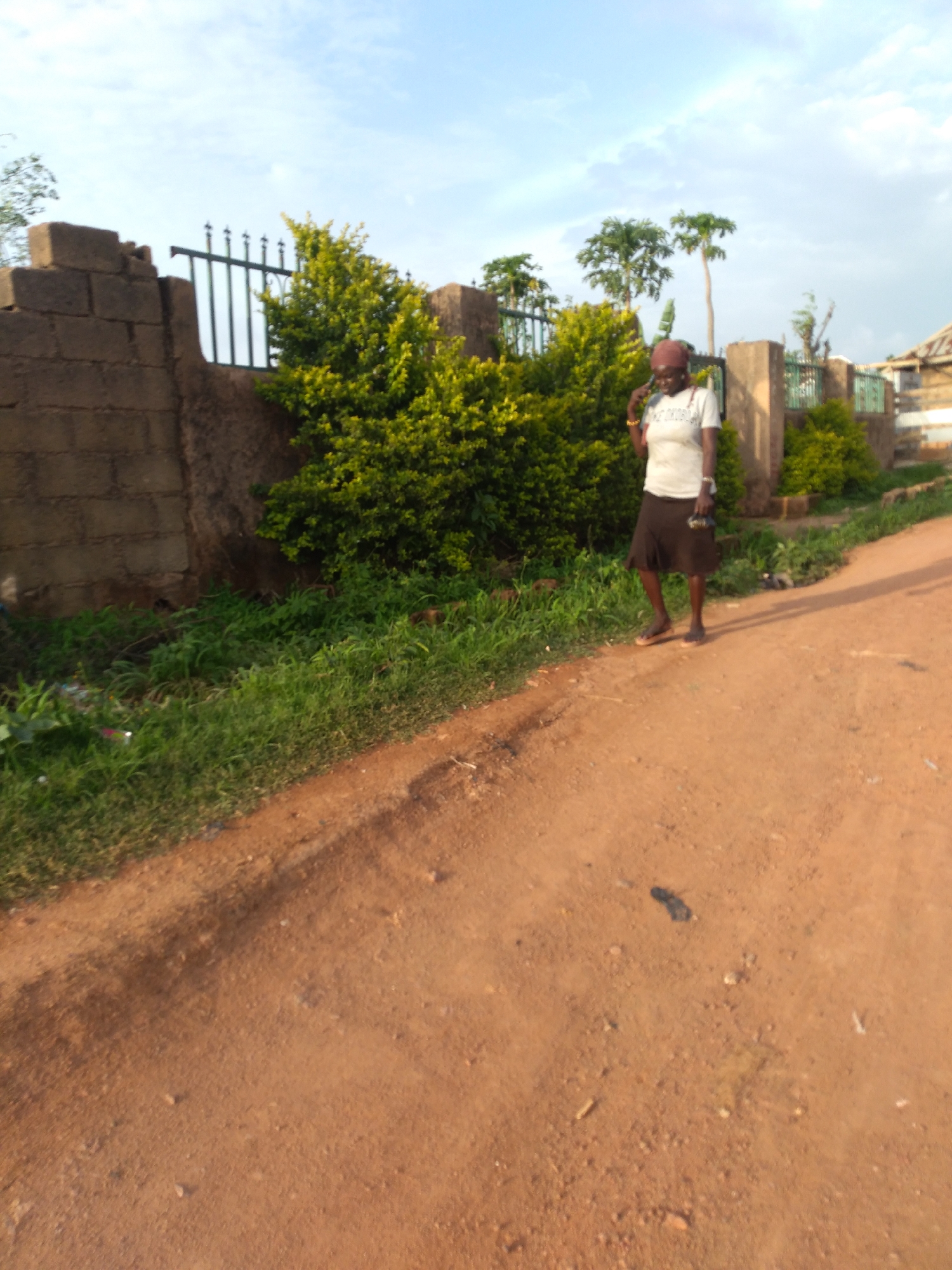 A woman is walking down a dirt road in a suburban area. She is wearing a white t-shirt, a brown skirt, and sandals. She is also wearing a brown headscarf. She is holding a phone to her ear. The road is lined with grass and bushes. In the background, there is a brick wall and a fence. The sky is blue with some clouds. The scene is lit by natural sunlight. The overall impression is one of a peaceful, everyday moment in a developing country. The location is implied to be Jos, Nigeria, a city in the north-central part of the country.