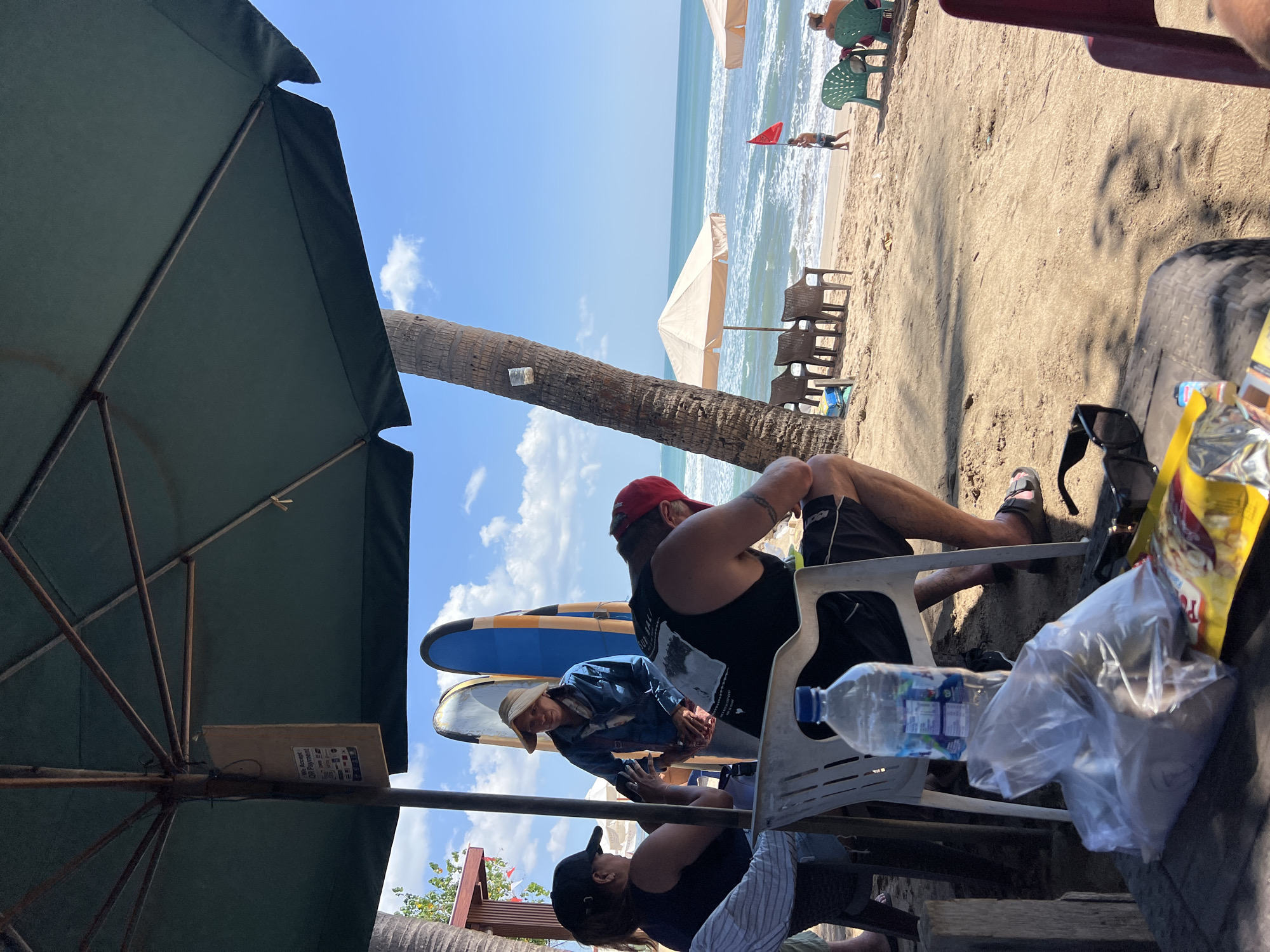 In Kuta, Indonesia, a beach scene unfolds under a large, dark green umbrella, casting shadows on the sandy ground. Two women and one man are seated at a table, partially obscured by the umbrella's support structure. One woman, wearing a black baseball cap and a dark sleeveless shirt, is holding a phone, seemingly taking a photo. The other woman, in a light-colored hat and a denim jacket, stands nearby, holding a phone and what appears to be food items. The man, in a black tank top and red baseball cap, is seated with his back to the camera, looking down. A plastic bottle of water and bagged food items are on the table in the foreground.
Beyond the seated individuals, the sandy beach stretches towards the ocean, dotted with chairs and beach umbrellas. Surfboards stand upright near the water's edge. In the distance, several people are visible on the beach, some sitting on chairs and one person walking near the water, holding a red flag. The sea has moderate waves, and the sky is blue with scattered clouds. The overall atmosphere suggests a relaxed beachside gathering or cafe.