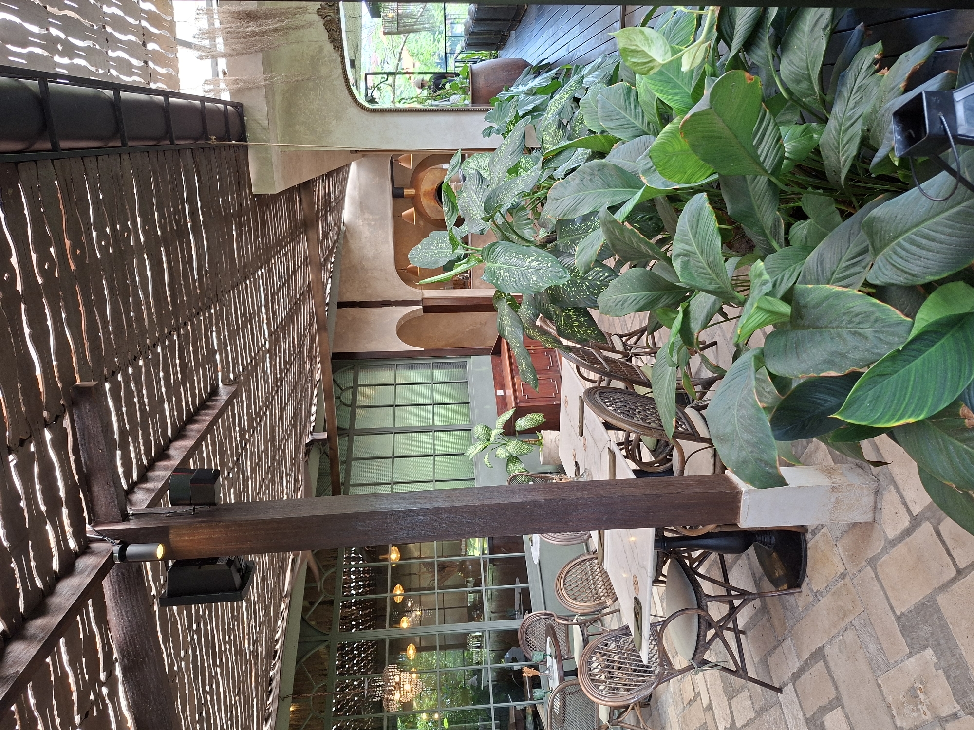 The image captures an outdoor dining space in Ubud District, Indonesia. The foreground is dominated by lush green plants, including peace lilies and dieffenbachia, beside woven chairs around tables with white marble tops. The tables are arranged on a brick-paved area. Overhead, a wooden pergola with an open slat roof structure filters the light. A dark-stained wooden support beam runs vertically, with lighting fixtures attached. In the background, large windows offer views to a garden or greenery outside, with a chandelier visible inside. The overall scene presents a relaxed, natural ambiance.