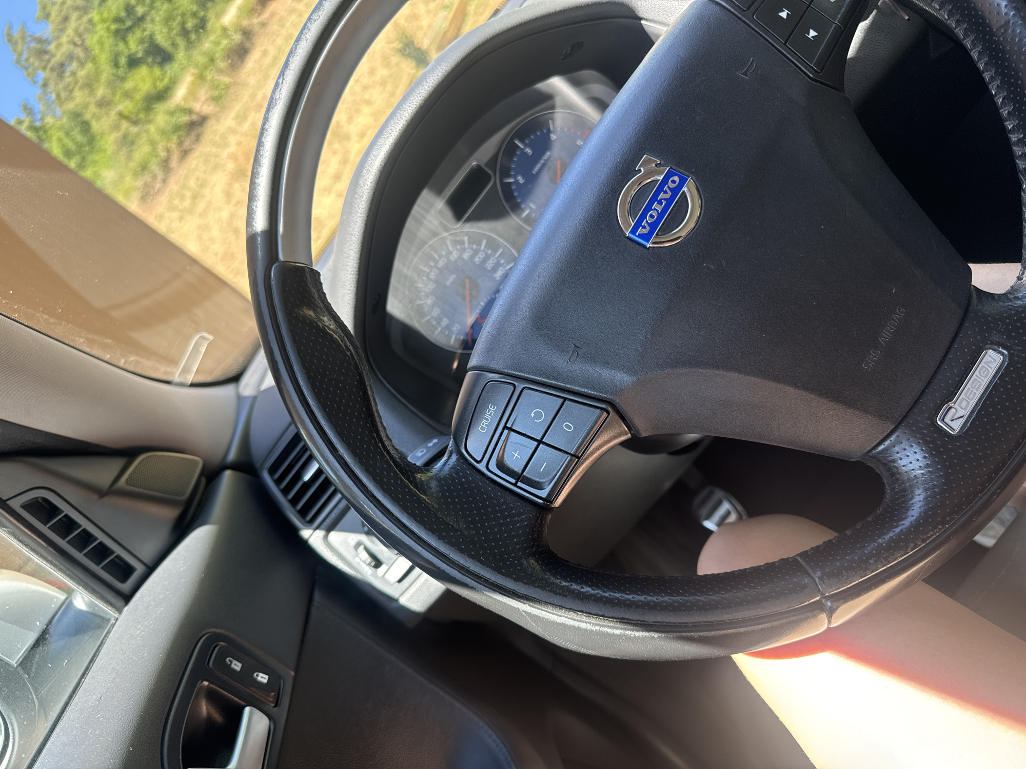 The image shows the interior of a Volvo car, focusing on the steering wheel and dashboard. The steering wheel is black leather with perforated grips and features controls for cruise control, volume, and other functions. The Volvo logo is prominently displayed in the center of the steering wheel. The dashboard includes a speedometer and tachometer. Through the windshield, a sunny, rural landscape with dry grass and green trees is visible. A person's bare leg is visible in the lower left corner of the image, suggesting the driver or a passenger is in the vehicle. There is no information in the image that allows for the determination of the location in the city of Saint-Claude, France.