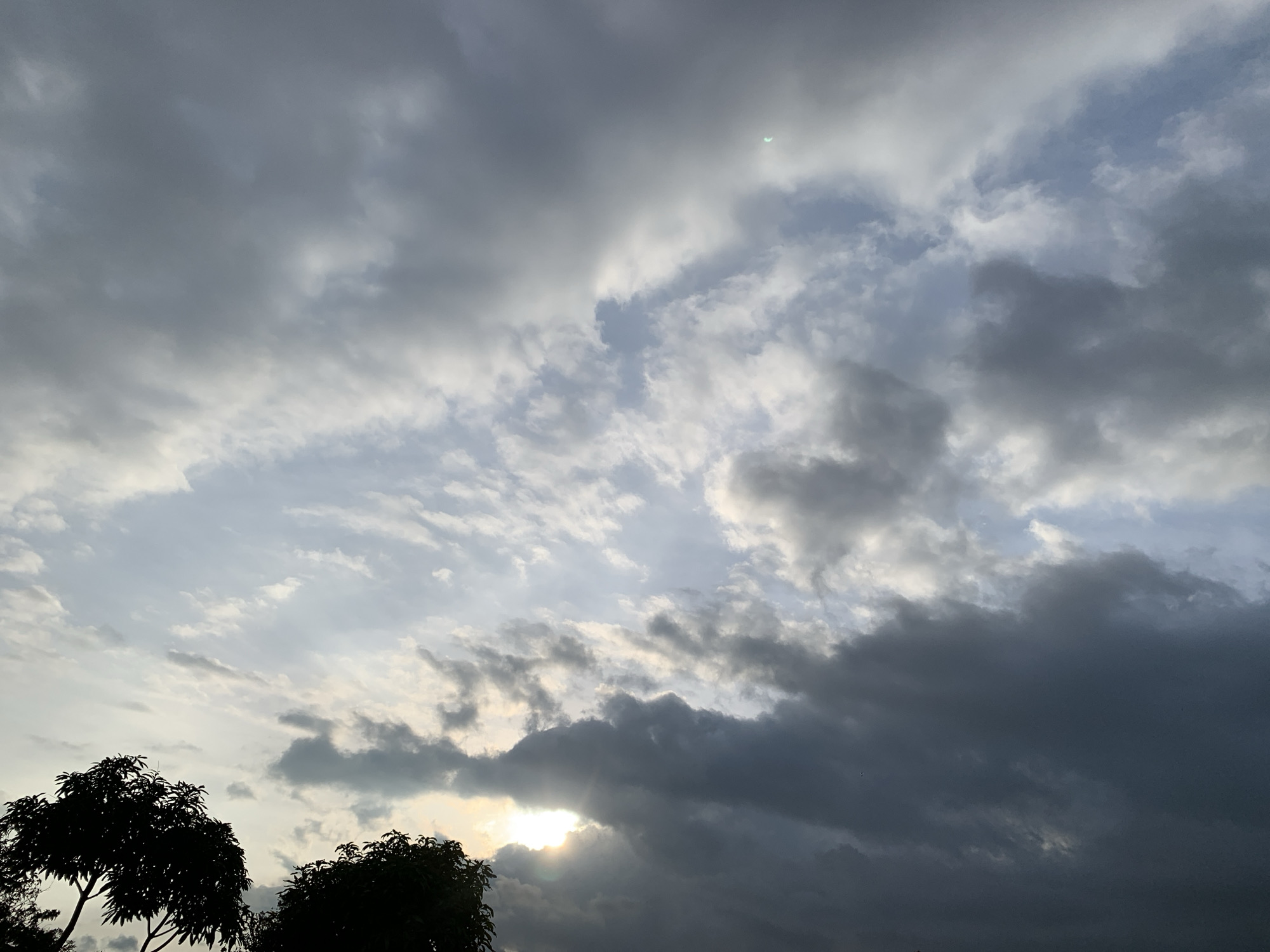 The image shows a dramatic sky filled with a mixture of dark, heavy clouds and lighter, wispy formations. Sunlight breaks through the clouds in places, creating bright highlights and casting shadows. In the lower left corner, the silhouettes of two trees are visible against the sky. The scene appears to be set in an open area, possibly on the outskirts of a city. The atmosphere is overcast, suggesting either the approach of rain or the aftermath of a storm. The location in Kuru Karama, Nigeria, is suggested by the mention of this Nigerian city.