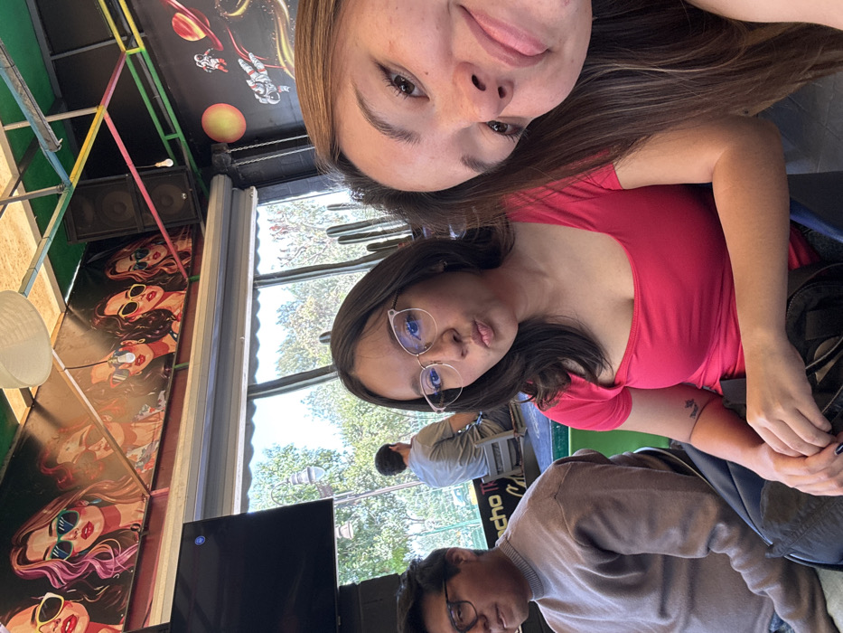 The image captures an indoor scene, likely a cafe or restaurant, located in San Andrés Cholula, Mexico, with an outdoor area visible through large windows. Three individuals are prominently featured indoors. In the immediate foreground, partially visible on the right, a female with long brown hair is taking a selfie, looking directly into the camera. To her left, a second female with dark hair and wearing glasses and a bright pink short-sleeved top makes a "pout" expression, also looking at the camera. To the left of this female, a male with dark hair and glasses wears a light brown turtleneck-style top and looks slightly to his left. The indoor space features decorative elements: the ceiling displays a white paper lantern-style light fixture and exposed beams, while the walls are adorned with pop-art murals depicting women in sunglasses and a separate space-themed mural with an astronaut and planets. A large black speaker is mounted on the wall. Through the expansive windows, an outdoor environment is visible, including tall, thin cactus plants, green trees, and a street light. A fourth male figure is seated outdoors at a table, seen from behind. A partial sign with the text "cho T" is visible outdoors. The scene is brightly illuminated.