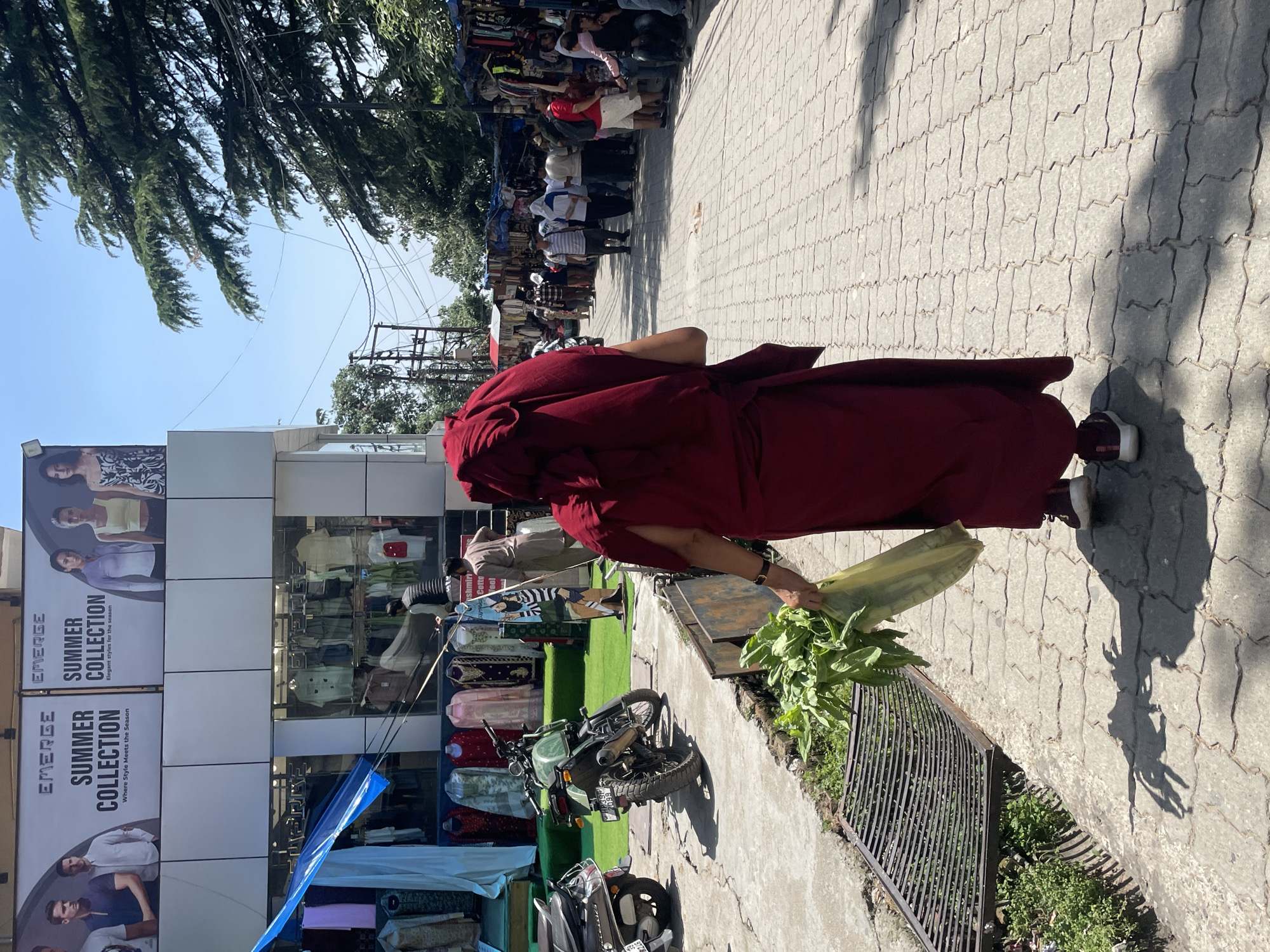 The image shows a person in a maroon robe walking on a paved sidewalk in Dharamshala, India. The person is carrying a bunch of green leafy vegetables. A row of shops lines the left side of the sidewalk, featuring the "EMERGE SUMMER COLLECTION" displayed on large advertisements. The shops appear to sell clothing and textiles, with some items displayed outside under a blue tarp. Several motorcycles are parked near the shops. Other pedestrians are visible in the background. The day is sunny, and the shadows are sharp. Trees can be seen overhead.