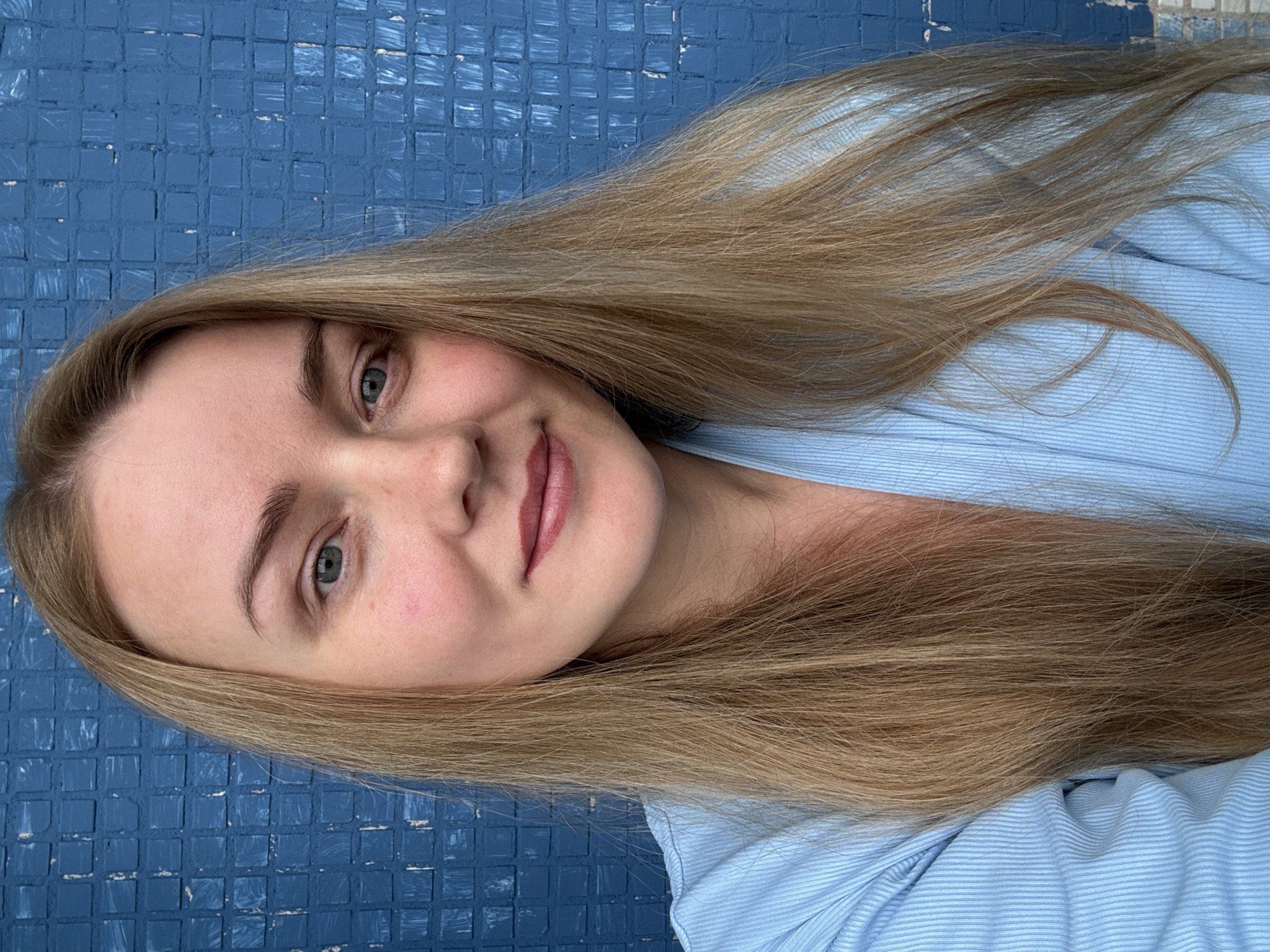 A medium-shot, eye-level, full-face portrait of a young Caucasian woman with long blonde hair, fair skin, blue eyes, and a light pink lipstick is featured against a textured blue wall. She is wearing a light blue, pinstriped collared shirt. The woman's head is tilted slightly to her left. The image is an outdoor candid shot, likely taken in Chaikovsky, Russia, given the blue tiled wall which is a common feature in Russian public spaces. The lighting suggests natural daylight.
