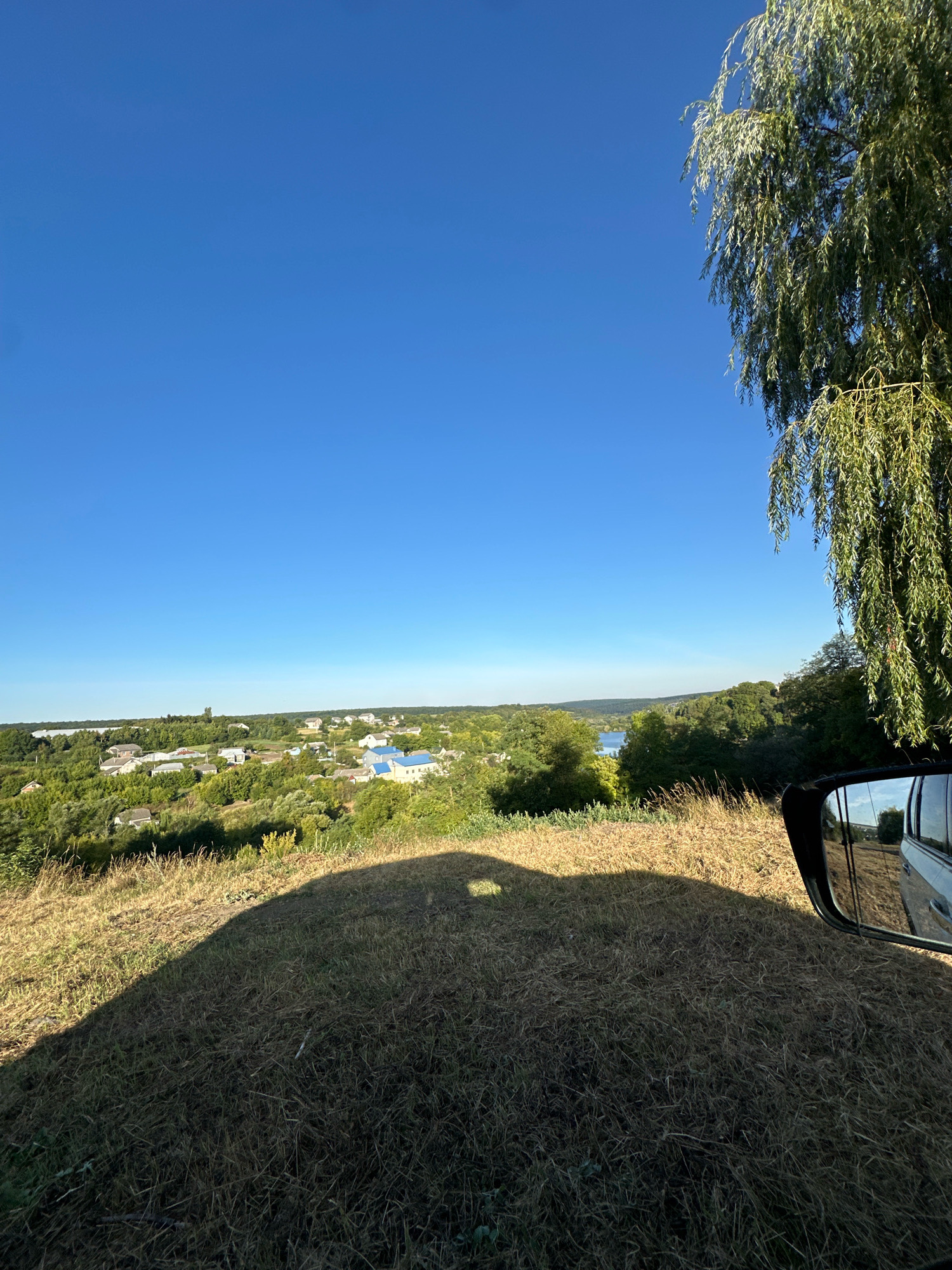 A wide-angle, outdoor, eye-level shot captures a rural landscape with a clear blue sky dominating the upper half. A large weeping willow tree is positioned in the upper right quadrant, its long, green branches extending downwards. In the middle ground, a village with houses, some with blue roofs, is nestled among lush green trees. Beyond the village, a body of water is visible, bordered by rolling green hills in the distance. The foreground is a grassy slope covered in dry, light-brown grass, with a large shadow of a vehicle cast across it from the left. A car's side mirror is visible on the right side of the frame, reflecting a rural road and trees. The scene suggests a sunny day in a tranquil, possibly Ukrainian, rural setting. The location is identified as Makiv, Ukraine, though this cannot be visually confirmed from the image itself.