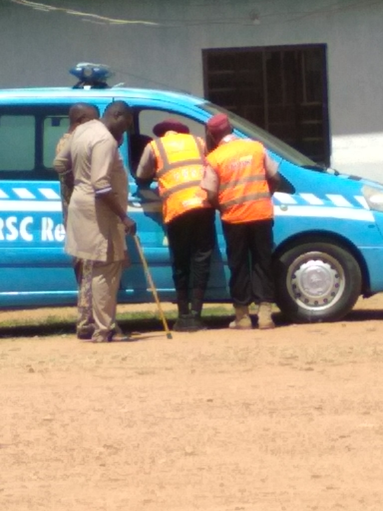 The image captures an outdoor scene in Kuru Dungus, Nigeria, during daylight hours under clear weather conditions. A light blue utility or rescue van is prominently featured, positioned on dusty, reddish-brown ground. The van is equipped with a blue light bar on its roof and has white stripe markings along its side. Partially visible text on the van reads "RSC Re," suggesting it may belong to an organization like the Federal Road Safety Corps (FRSC Rescue).
Several individuals are gathered around the driver's side of the van. One man, dressed in a light-colored traditional tunic and trousers, stands holding a wooden walking stick, facing the side of the vehicle. Behind him, another person is partially visible. Two other men, seen from behind, are wearing bright orange reflective safety vests over dark attire, indicating they might be part of an official or emergency service. One of these men also wears a maroon beret. They appear to be engaged in an activity involving the van, possibly attending to someone or something inside or near its open door.
In the background, a simple, light-colored building with a large, dark rectangular opening, possibly a window or doorway, is visible. The overall scene suggests a roadside stop or an incident response in a public or semi-public area.