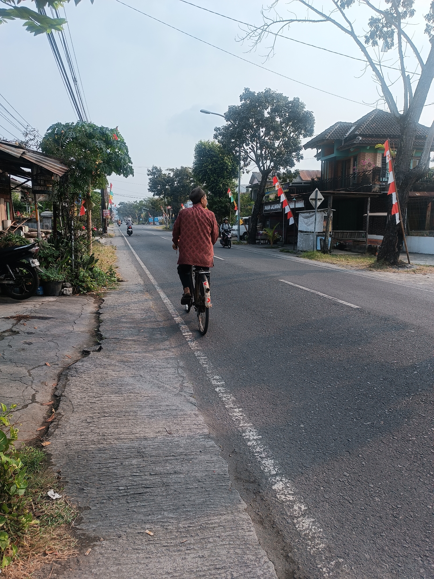 A person is riding a bicycle on a paved road in Sewon, Indonesia. The individual is seen from behind, wearing a patterned shirt and dark pants, pedaling away from the camera. The road is asphalt with a white line marking the edge of the lane. To the left of the road, there is a concrete sidewalk with some vegetation and a parked motorcycle. Along the sidewalk are several small buildings or stalls, some decorated with red and white flags, suggesting a festive occasion. Trees line the road, and in the background, more buildings and trees are visible, with a light-colored sky overhead. Several other motorcycles are also seen on the road in the distance.