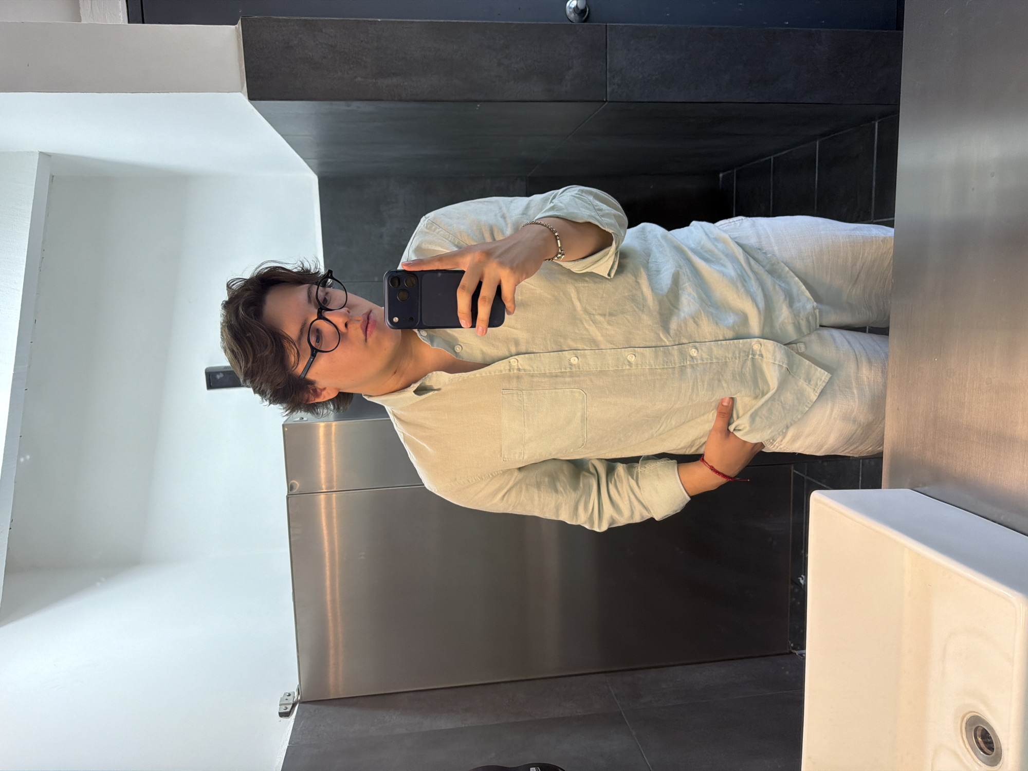 A person is taking a selfie in a bathroom, likely in Puebla, Mexico. The individual is wearing glasses and a light-colored button-down shirt and pants. They are holding a dark-colored smartphone in their right hand, presumably to take the photo in the mirror. The bathroom has a white sink and stainless steel fixtures. The wall behind the person is dark gray, possibly tiled.