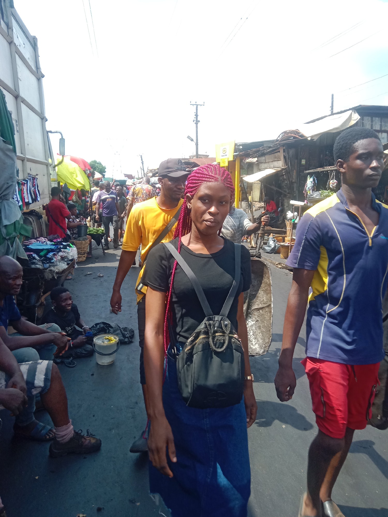 This vertical full shot captures a bustling, sunlit street market scene in Onitsha, Nigeria. In the immediate foreground, a woman with striking reddish-pink braided hair, wearing a black t-shirt, blue denim skirt, and a black backpack, walks towards the viewer with a neutral expression. To her right, a man in a blue and yellow polo shirt and red shorts also walks forward, partially visible. To the left, a man in a blue shirt and striped shorts is seated on the ground.
The midground depicts a lively market with numerous people moving between makeshift stalls. Behind the woman, another man in a yellow t-shirt and dark baseball cap walks past. On the left, a large white truck or van is parked beside stalls displaying clothing items. A child is seated on the ground near a white bucket, smiling, while another person operates a pushcart further back. Market stalls with various goods, some covered by tarps, line both sides of the crowded thoroughfare, indicating active commerce.
The background shows more market structures, people, and utility poles. The sky is bright and clear, suggesting sunny weather during the day. Overhead power lines are visible across the frame. A yellow sign bearing the "MTN" logo, a mobile network provider, is discernible in the mid-background, confirming a commercial presence. The ground appears to be a paved or packed surface. The overall scene conveys the vibrant energy of a typical African open-air market.
