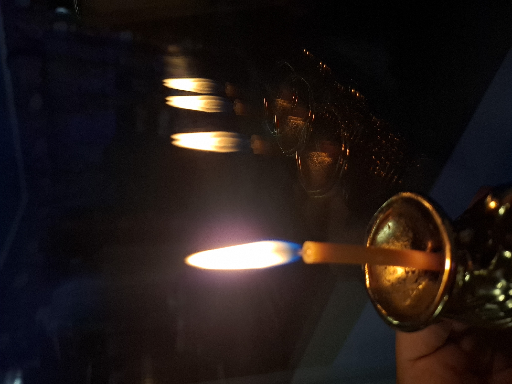 A lit candle in a metallic holder is held by a person in a dark environment. The flame is elongated, and its reflection appears several times in the background. The location is not discernible in this image.
