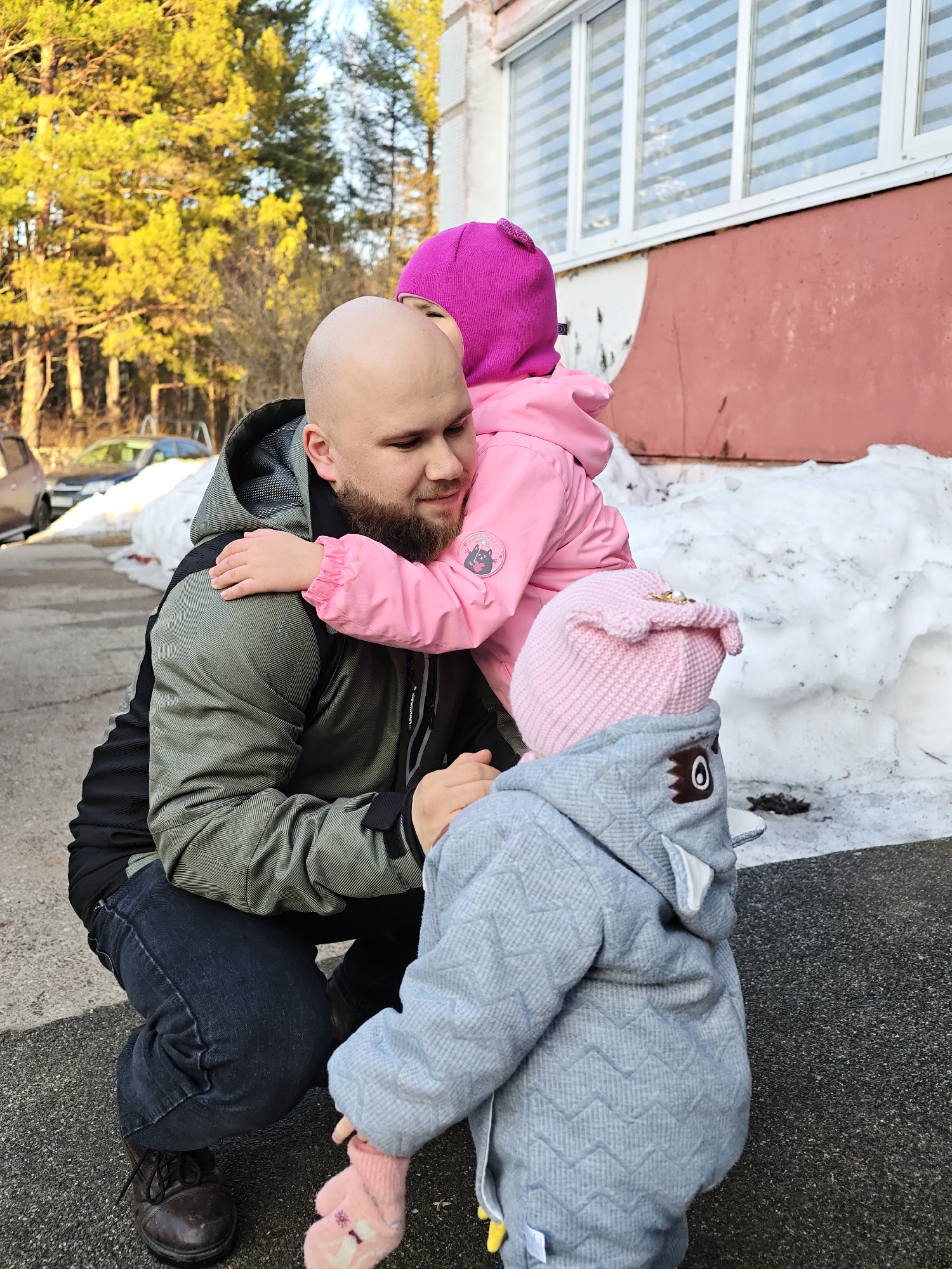 An outdoor scene in Chaikovsky, Russia, depicts an adult male interacting with two young children. A bald, bearded male, wearing a green and black patterned hooded jacket, black undershirt, dark blue jeans, and brown lace-up shoes, is crouched on dark pavement. He is looking down with a gentle expression as a child in a pink hooded jacket with a round patch and a fuchsia knitted hat hugs his neck. A second child, partially visible in the foreground and facing away from the camera, wears a light blue textured coat with a hood designed to resemble an animal (featuring a brown eye patch and white ears), a pink knitted hat, and pink mittens. Patches of dirty snow are visible on the ground to the right and behind the subjects. In the background, a light-colored vehicle and a dark car are partially obscured by snow. A building with a red lower section and a white upper facade, featuring a row of windows with horizontal blinds, is visible to the right. Beyond, a dense stand of coniferous trees with yellowish-green needles, illuminated by sunlight, rises against a clear light blue sky.