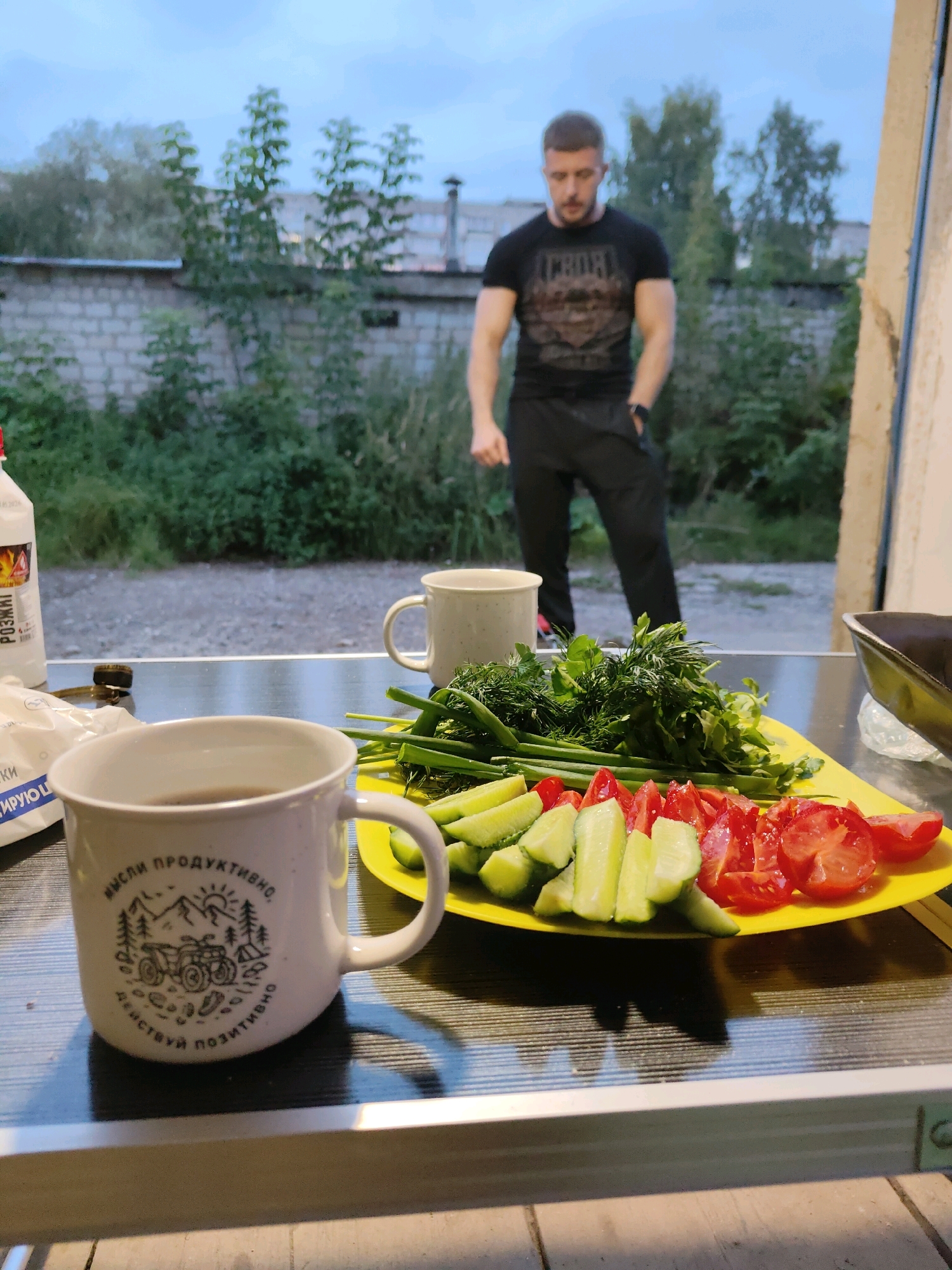 A man stands outside a building with a brick wall in the background, a hazy overcast sky above. In the foreground, a table is set with two mugs, one with dark liquid, and a yellow plate piled with chopped tomatoes, cucumbers, green onions, and dill. A bottle of lighter fluid and a bag of napkins are also on the table. The man, dressed in a black t-shirt and black pants, appears to be looking down. The scene suggests a casual outdoor meal or gathering.