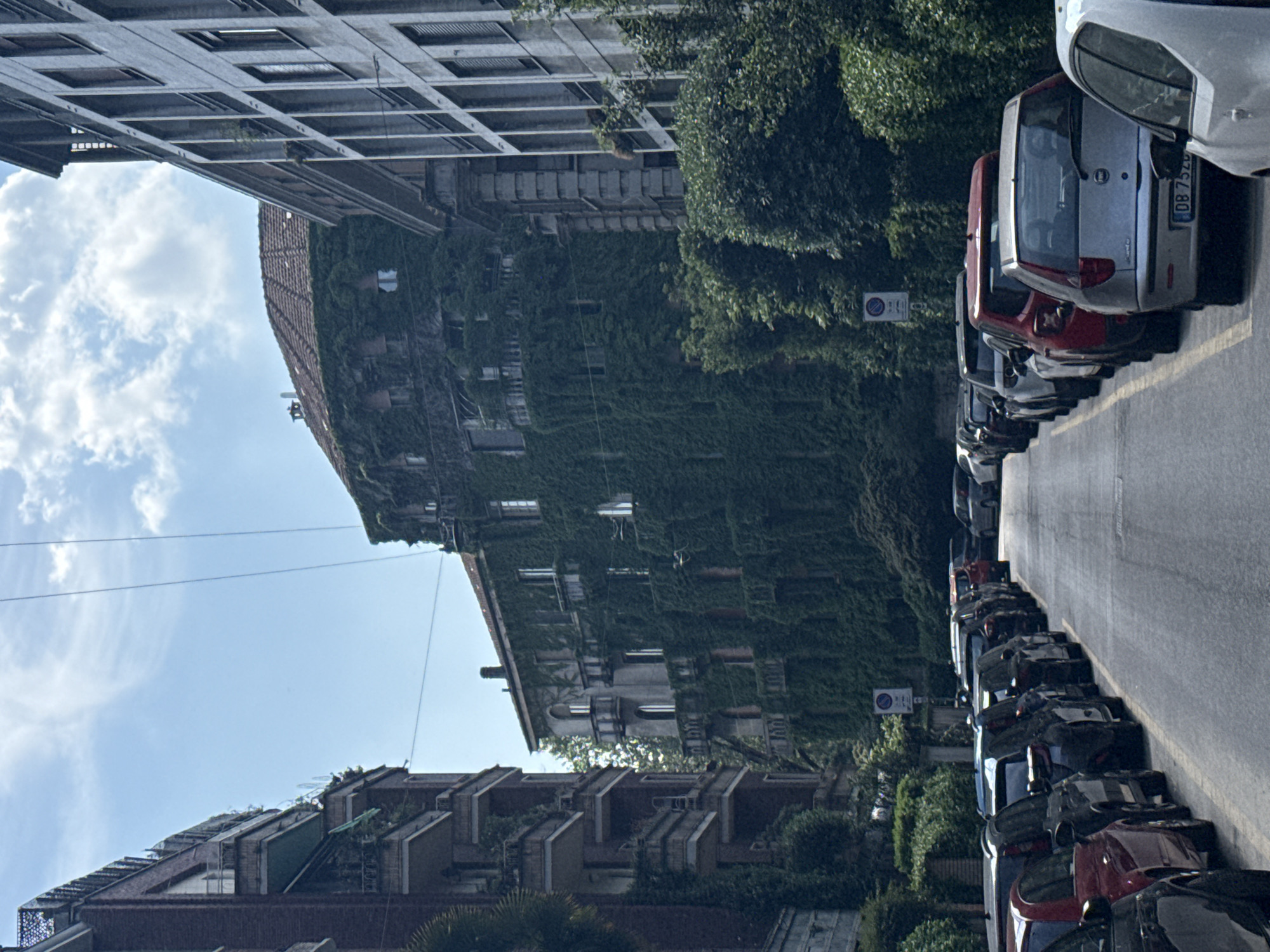 The image captures a vibrant daytime street scene in an urban environment, likely Milan, Italy, as indicated by the context. The photograph is oriented vertically, showing a street lined with buildings and parked cars under a clear sky with scattered clouds.
In the foreground, a row of cars is parked bumper-to-bumper along the right side of a paved street. The vehicles are diverse in model and color, with one car's license plate partially visible as "DB 752...", consistent with Italian registration plates. The asphalt road surface is visible to the left of the parked cars, marked by a yellow line. Several circular blue and white street signs, likely indicating parking regulations, are interspersed among the trees lining the street.
The midground is dominated by a distinctive building almost entirely enveloped in dense green ivy or climbing plants, with windows peering through the foliage. To its left, a taller, modern building features a geometric, grid-patterned facade, possibly made of concrete or stone. Further down the street, on the left side, another building with a terraced or stepped roof design, suggesting balconies or roof gardens, adds to the architectural variety.
The sky above is bright blue with scattered, wispy white clouds, indicating fair weather. Lush green trees are visible along the street, complementing the ivy-covered building and contributing to the urban greenery. Thin utility lines can be faintly seen stretched across the sky between the structures. The scene is calm and characteristic of a typical city street during the day, with no people visible, focusing instead on the architectural and natural elements.