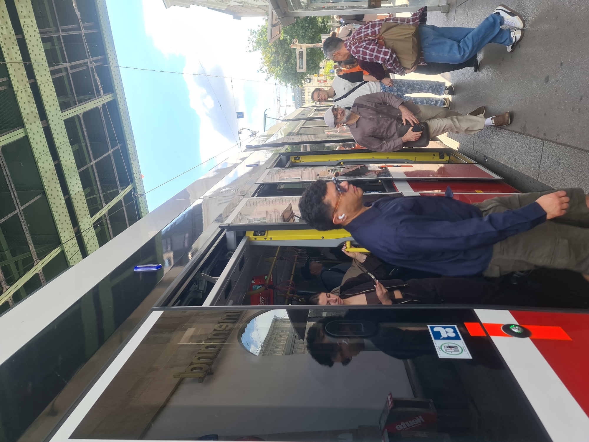 A group of people are standing on a street platform next to a tram. The tram doors are open, and people are entering and exiting. The tram is black and red, and the doors are yellow. The street platform is gray and there are trees and buildings in the background. The sky is blue with some clouds. The image appears to have been taken in Vienna, Austria, given the presence of a tram which is a common mode of transportation there, and the architectural style of the buildings visible in the background, which are characteristic of Viennese architecture. In the foreground, a man in a blue shirt is looking down, while a woman in a brown jacket is looking at her phone. A man in a brown shirt and a gray hat is standing next to her. Behind them, a man in a white shirt and glasses is looking forward. A man in a plaid shirt and jeans is standing to the right of the man in the white shirt.