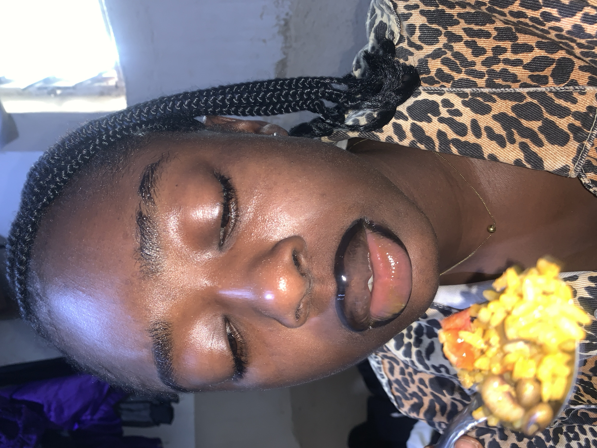 A close-up, high-angle shot shows a young Black woman with her eyes closed, about to eat from a spoon. The woman has dark skin, braided hair, and is wearing a leopard-print jacket and a thin gold chain. The spoon contains rice, beans, and what appears to be tomatoes. The background is out of focus and shows a wall and a window with bright light filtering through. The scene is indoors, likely in a residential setting in Rafin Par, Nigeria.