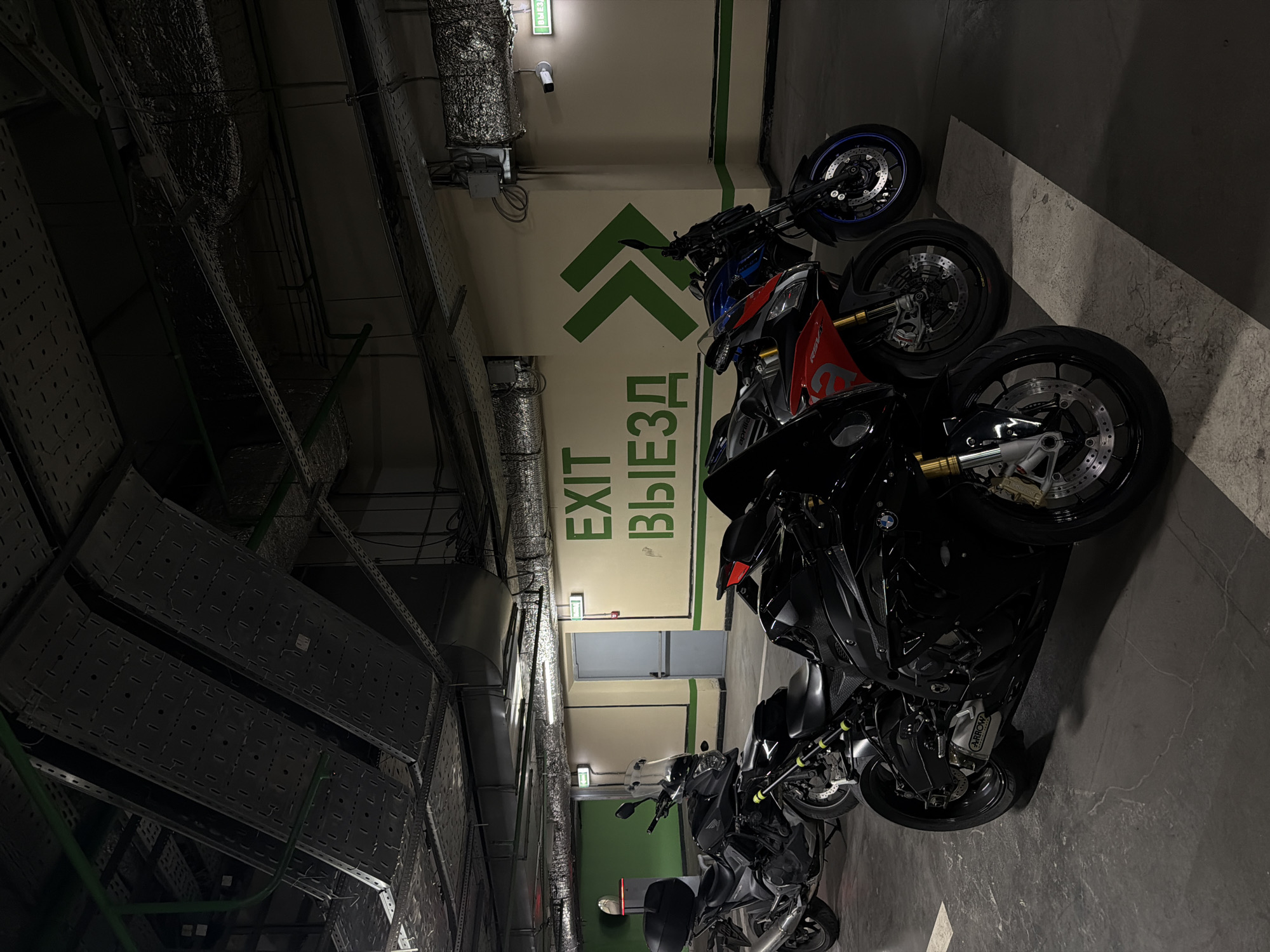 The image depicts a parking garage interior, likely in Moscow, Russia, given the presence of the Cyrillic word "ВЫЕЗД" (VYIEZD), meaning "EXIT," alongside the English word "EXIT" on a green sign with upward-pointing arrows. The sign is mounted on a wall within the garage. Several motorcycles are parked in a row. They include a BMW S1000RR with an Arrow exhaust system, an Aprilia RSV4, and a blue motorcycle with a single-sided swingarm. The surrounding environment consists of the parking garage's ceiling, which features extensive ventilation systems and ductwork. The floor is concrete. The overall lighting in the scene is dim.