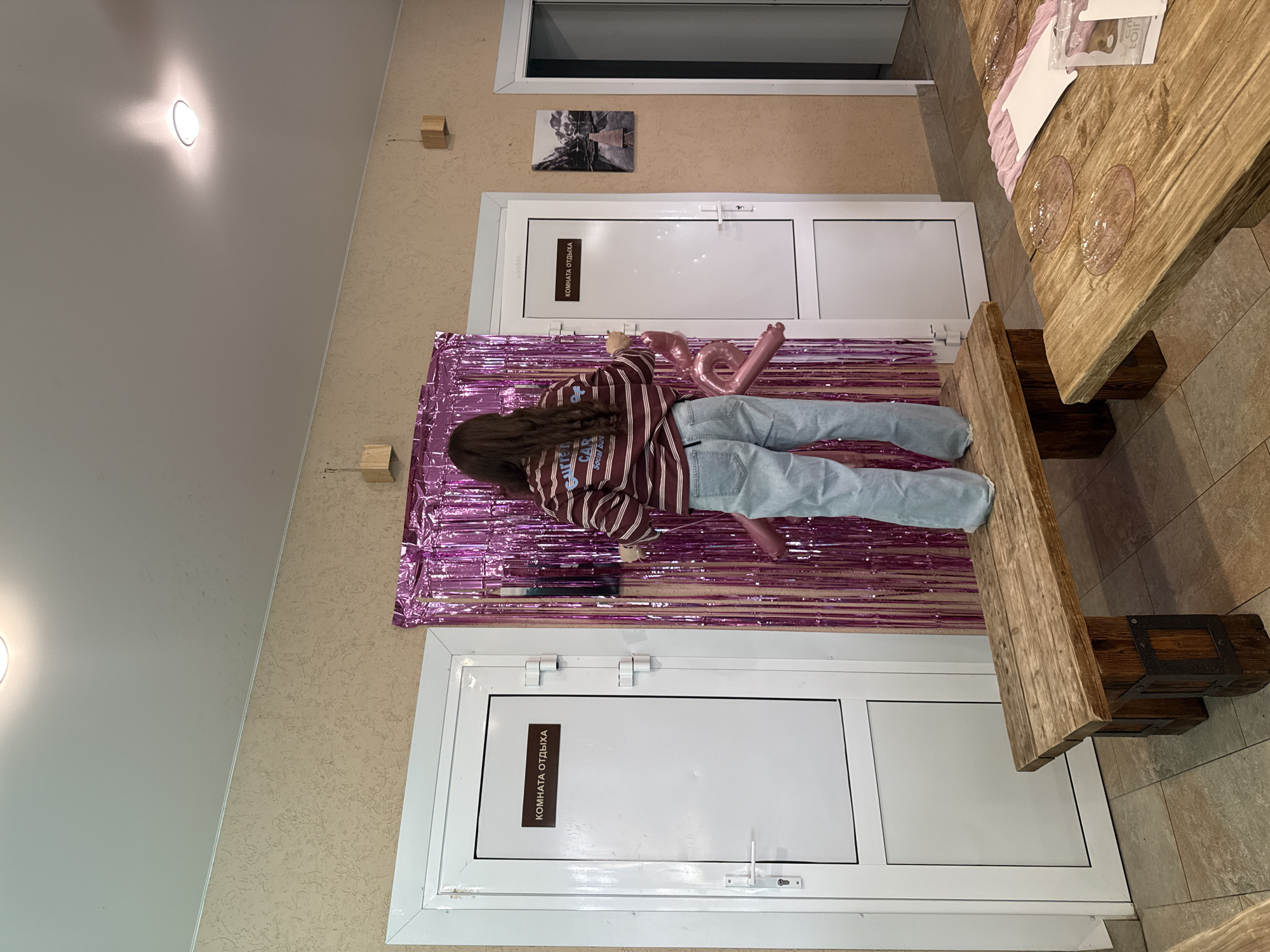 A young woman with long dark hair is lying on the floor in front of a purple tinsel curtain. She is wearing a striped long-sleeved shirt and light blue jeans. Two pink balloons, shaped like the numbers "1" and "6", are floating near her head. The background shows two white doors. One door has a sign that reads "КОМНАТА ОТДЫХА" (Rest Room). There is a small framed picture on the wall above this door. The scene appears to be indoors, possibly a party or celebration, given the balloons. The lighting suggests it is daytime or well-lit artificial lighting. Wooden tables with chairs are visible in the background, one of which has a few pink and clear glasses on it. The walls have a textured finish, and the floor is tiled.
